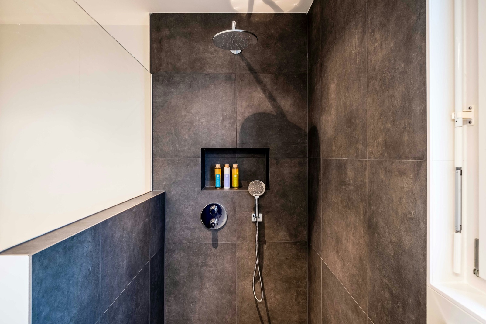 A walk-in shower lined with large dark grey stone-look tiles laid in a grid with thin matching joints, a rectangular niche set into the back wall holding three bottles, a chrome rain shower, a round concealed mixer and a hand shower, with a tiled half-height wall on the left