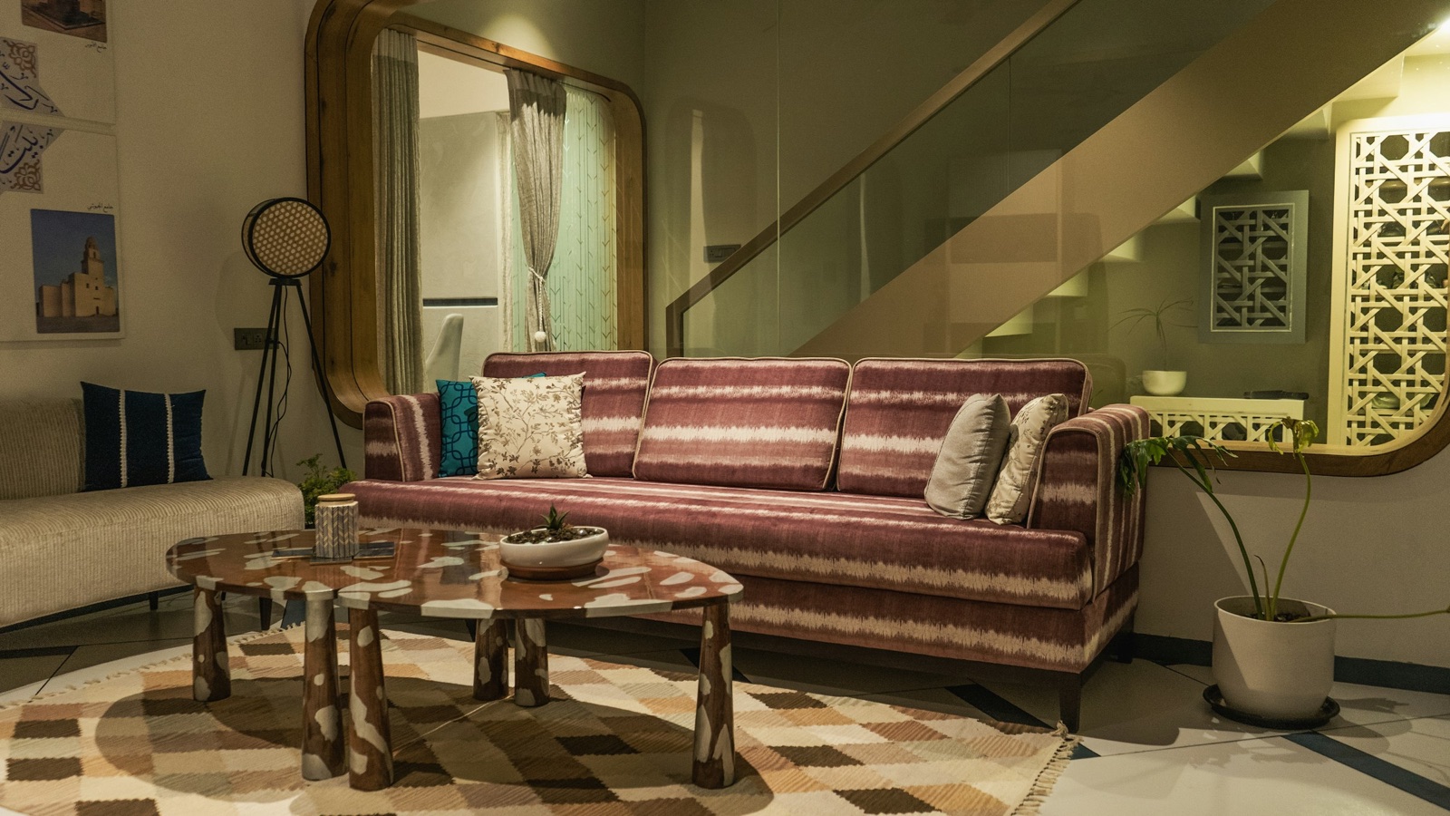 A three-seater sofa upholstered in a maroon and white striped woven fabric, on slim dark legs with narrow arms, beside a cream bench sofa, with a terrazzo-patterned coffee table on a checked rug, a glass-railed staircase and a carved white jaali screen behind