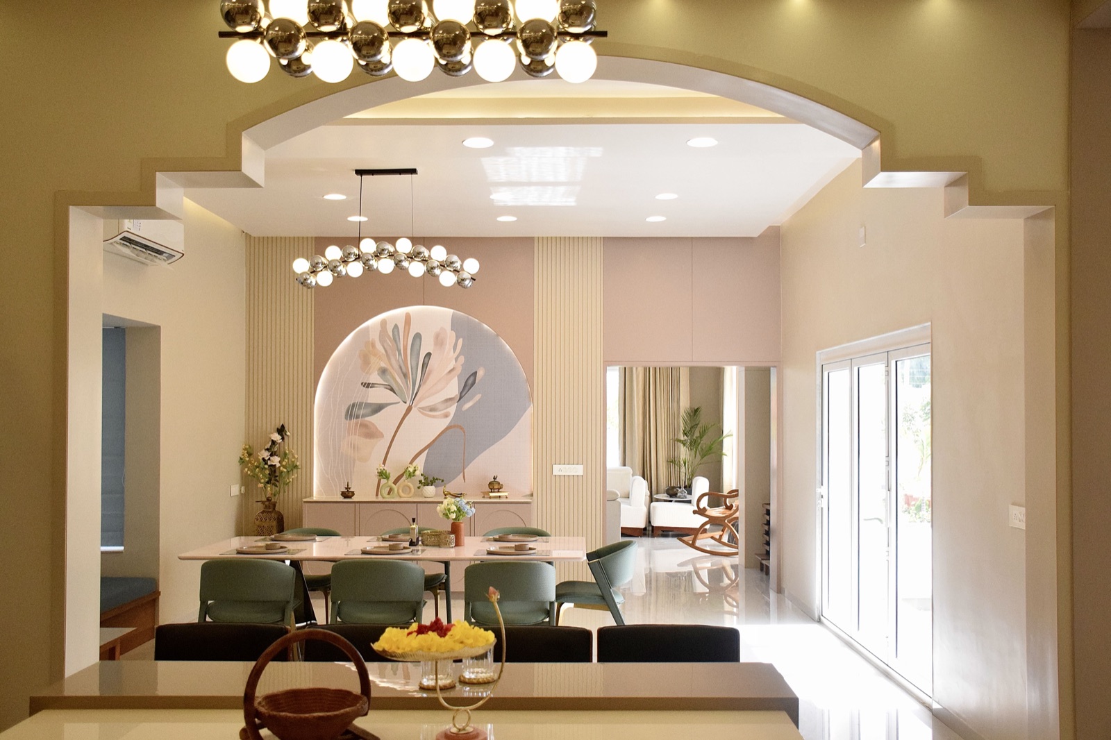View through an arched opening into a dining area with a long white table, sage-green upholstered dining chairs with curved backs, a sideboard under a lit arched niche with a painted leaf mural, fluted panels, a bubble chandelier, and a living room with a wooden rocking chair beyond
