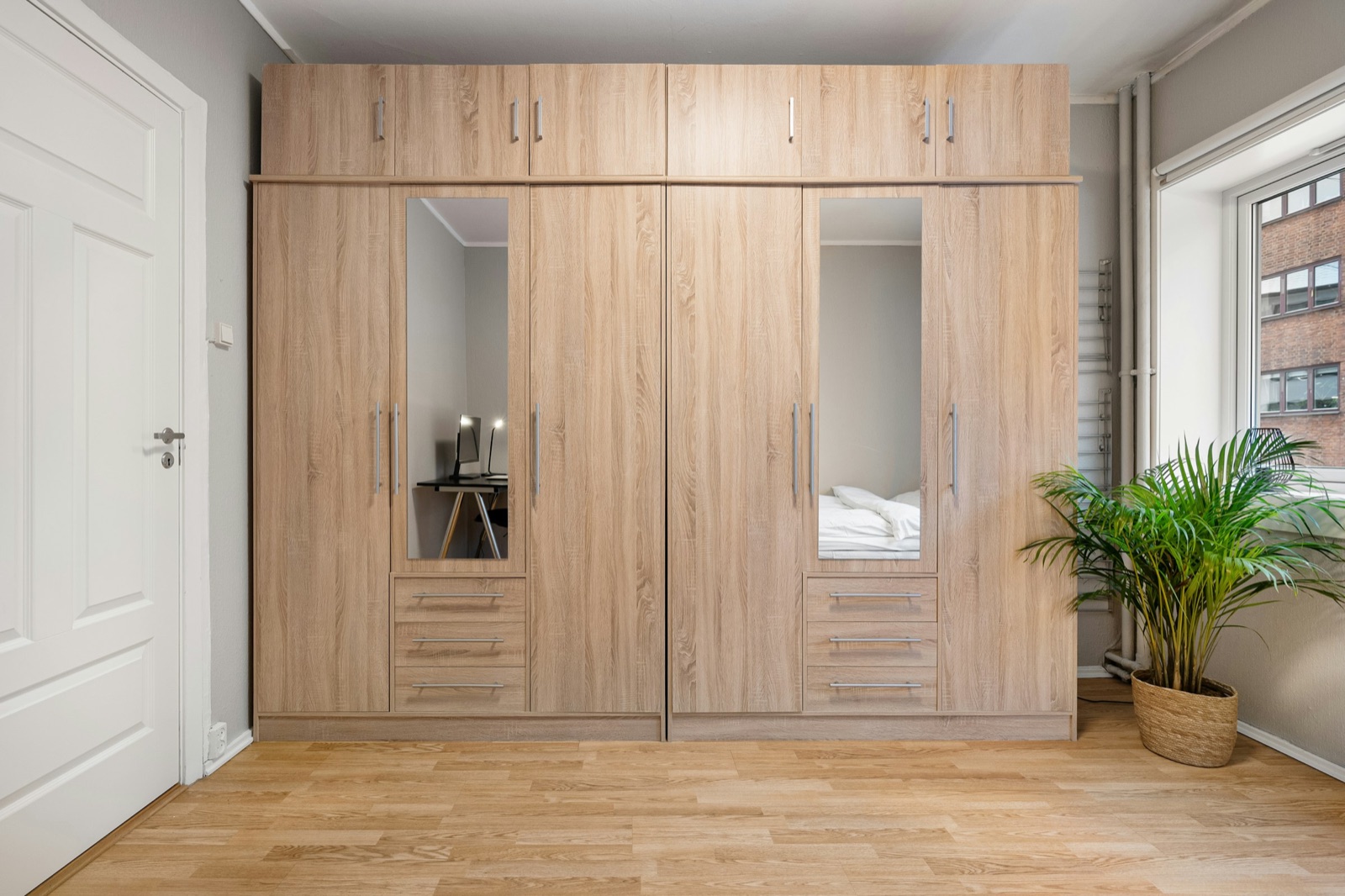 Two wardrobes side by side in a light oak wood-grain finish, each with a mirror panel in one shutter, three drawers below the mirror and a row of loft cabinets above, in a bedroom with a light wood floor and a white panelled door