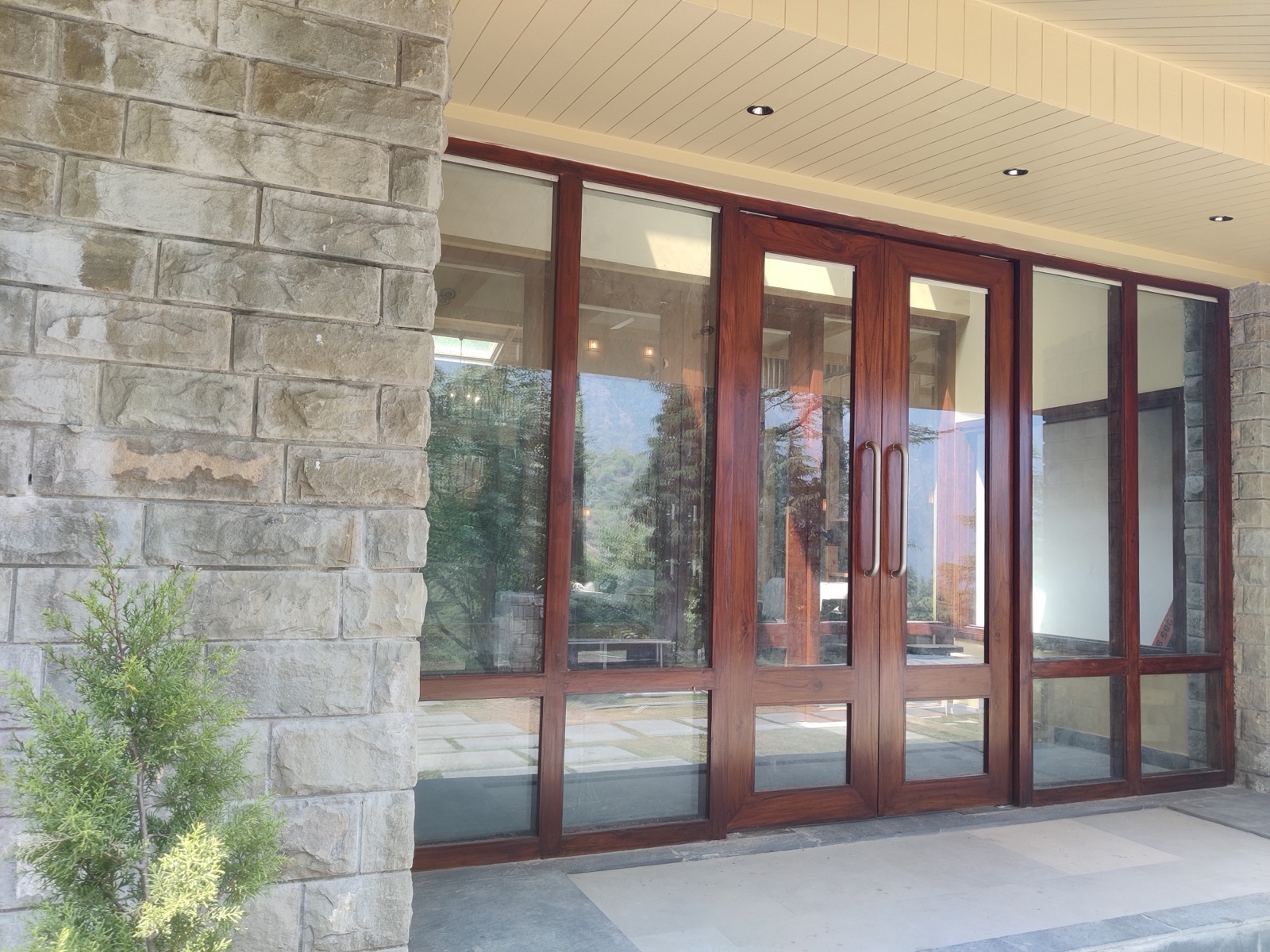 Entrance of a building in Dharamsala: a pier of large grey dressed-stone blocks laid in courses stands beside a wide glazed front with dark wood-toned frames and double doors, under a light panelled soffit, with stone-paved flooring and a small conifer in front.