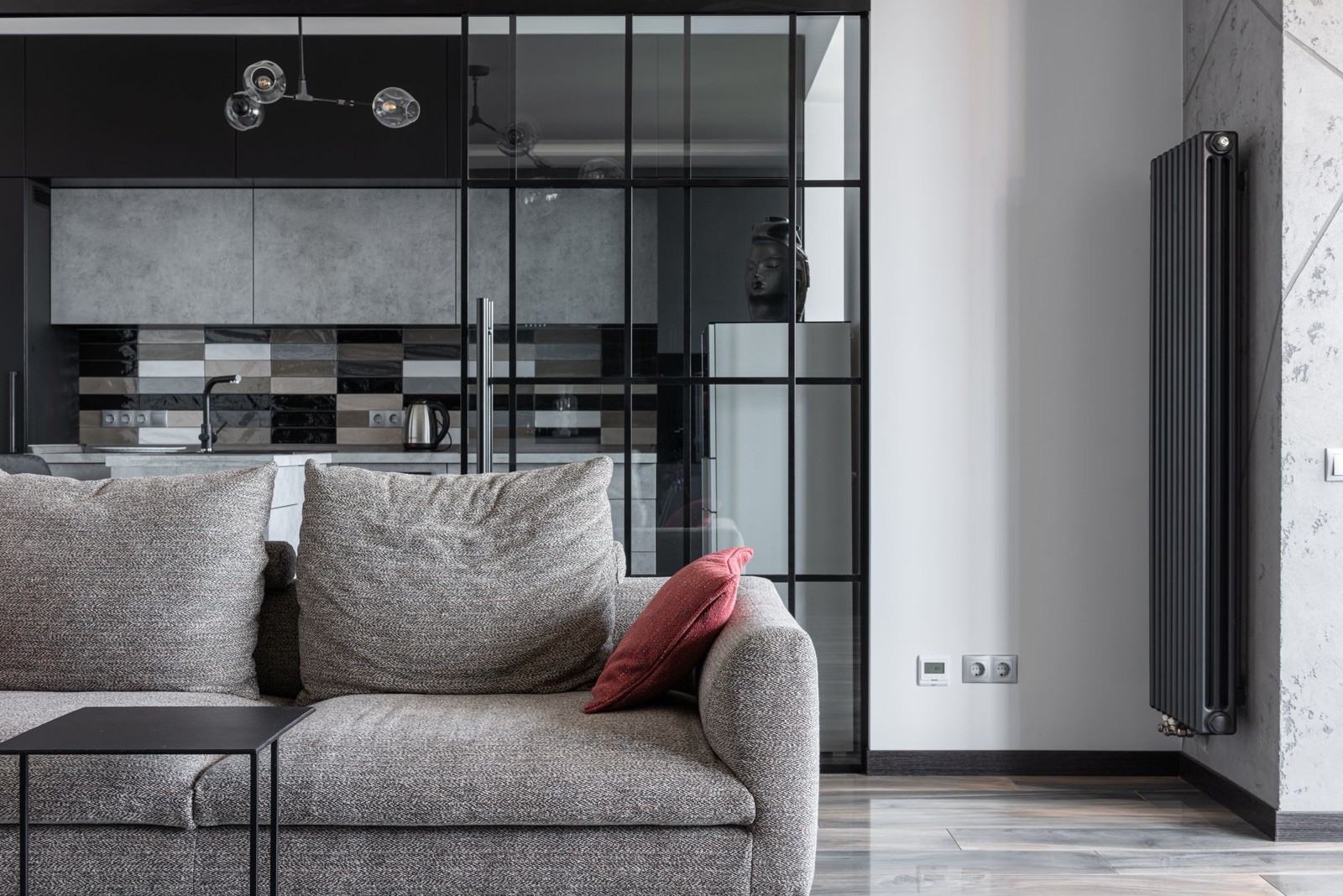 Grey fabric sofa with a red cushion in front of a full-height black-framed glass partition with a sliding door, through which a kitchen with grey wall units, a patterned tile backsplash, a sink and a kettle is visible.