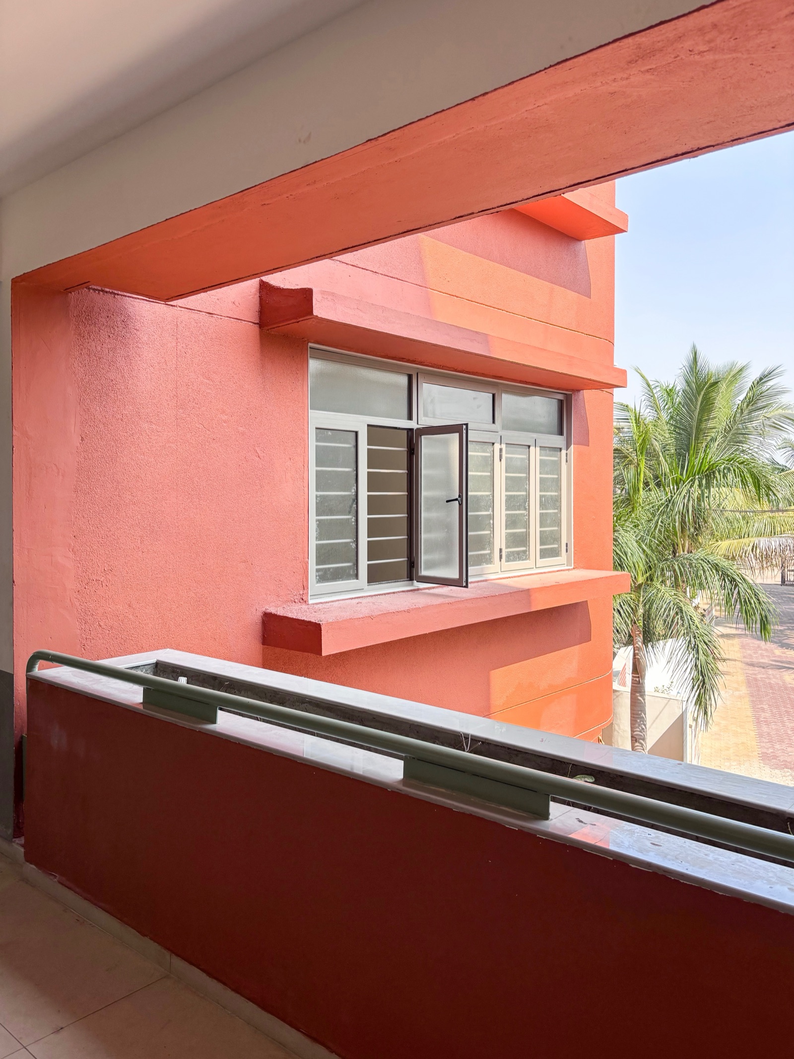 Terracotta-red exterior walls with a coarse grainy finish around a window with a projecting ledge, seen from a corridor with a deep red parapet, palm trees beyond