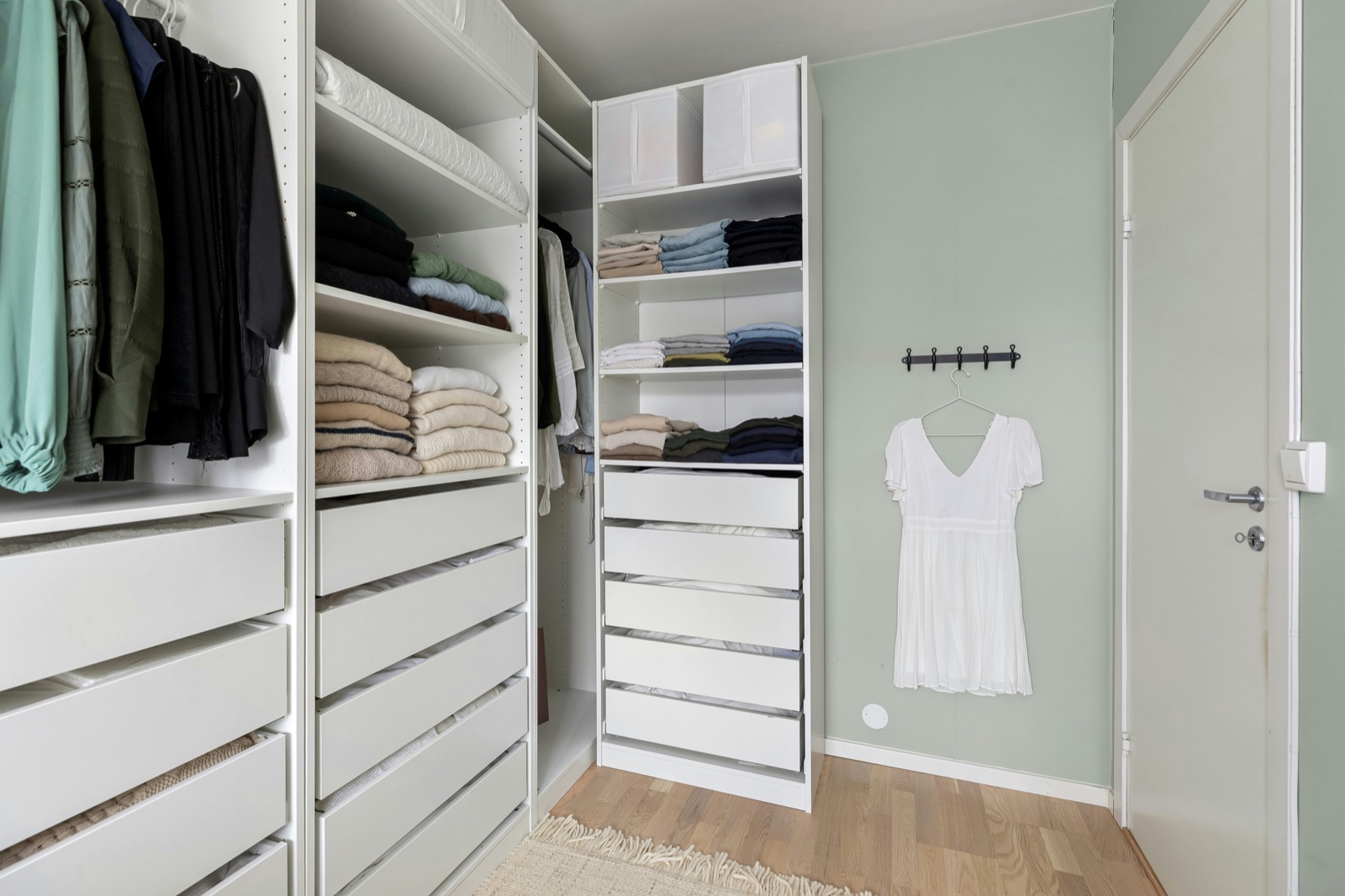 Open white modular wardrobe with long and short hanging clothes, shelves of folded sweaters and T-shirts in neat stacks, a column of five white drawers and storage boxes on the top shelf.