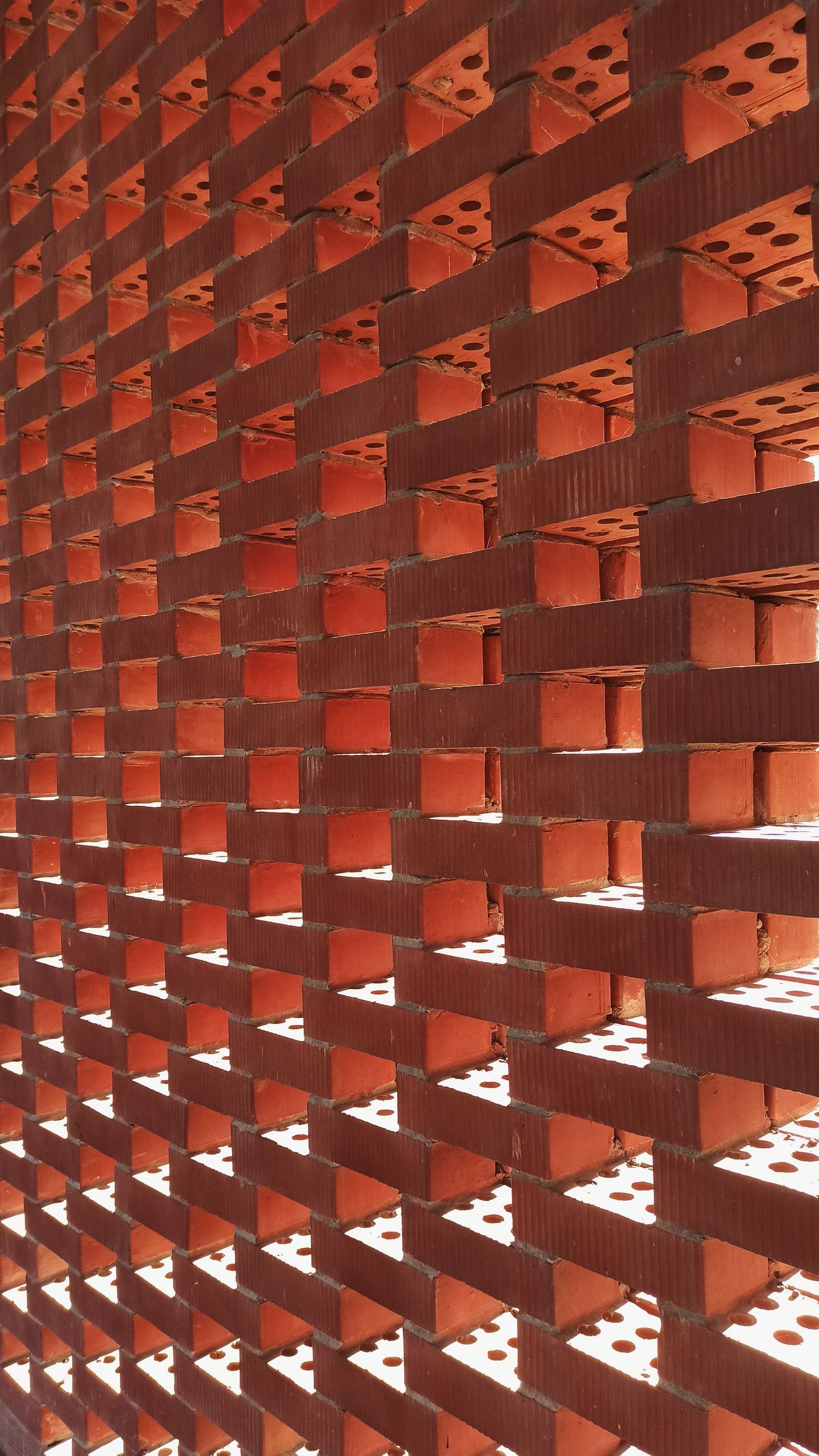 Close view of a red brick jaali wall: bricks laid in staggered courses with a gap between each pair, so bright daylight shows through rows of rectangular openings and the perforated bed faces of the bricks are visible inside each gap.