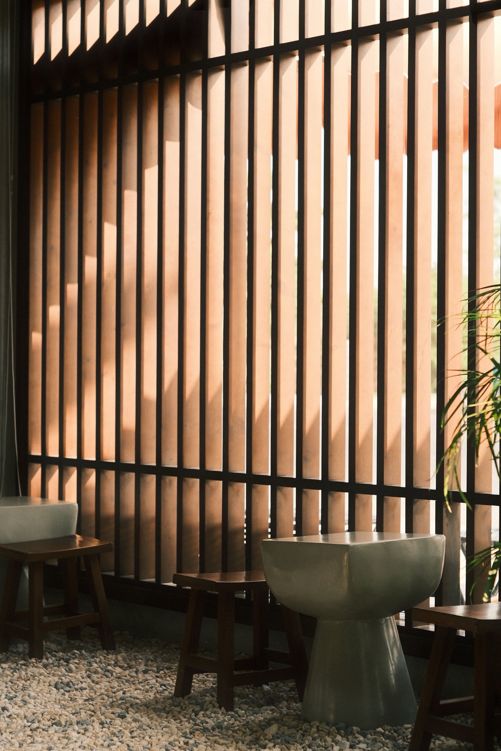 Close view of a floor-to-ceiling screen of square wooden battens held in a dark metal frame with horizontal rails, warm sunlight falling through the gaps onto the wood, with two wooden stools, a pale grey side table, a palm frond and a pebble floor in front