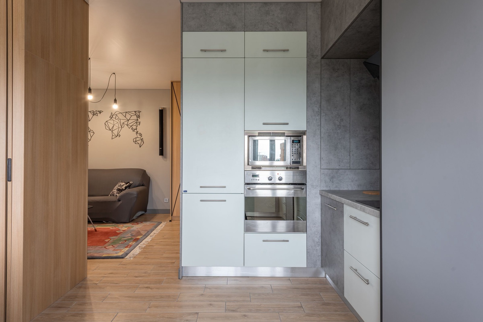 Tall bank of pale grey-green units at the end of a kitchen: a full-height two-door unit on the left and, on the right, a stainless steel built-in microwave stacked above a built-in oven, with a drawer below and cupboards above, and the living room visible through the doorway.