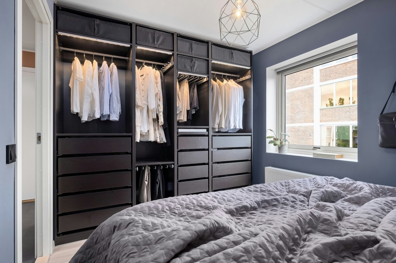 Bedroom with a wall of dark brown modular wardrobe frames without doors, four bays with lit hanging rails, storage boxes at the top, stacks of drawers and a pull-out trouser rail, taken almost to the ceiling beside a window in a slate-blue wall