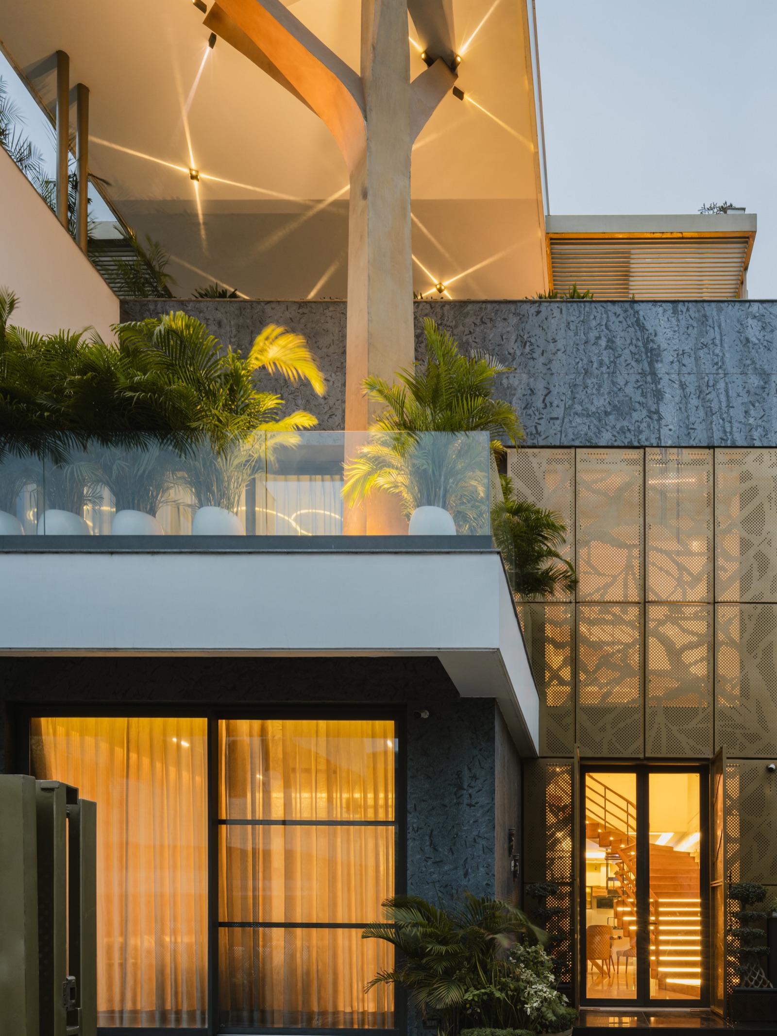 Dusk view of a house entrance where a pair of black-framed glazed doors stand open between gold perforated metal screen panels, with a lit staircase visible inside and dark stone cladding beside