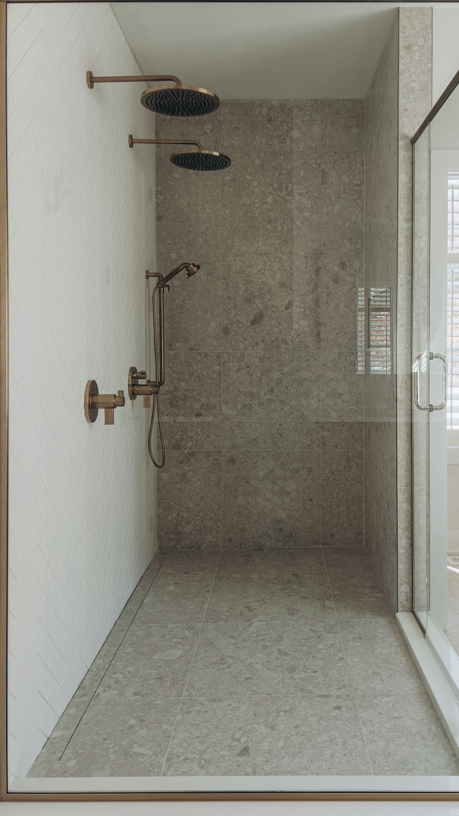 A walk-in shower seen through its glass door: large grey terrazzo-look tiles cover the floor and back wall, white herringbone tiles line the left wall, two brushed-bronze rain showers and a hand shower are fixed to the left, and a slim linear drain runs along the foot of the left wall