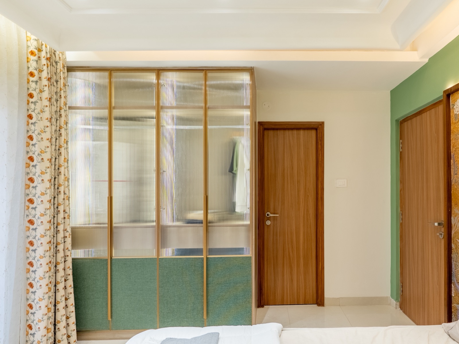 A bedroom wardrobe with four reeded glass shutters in thin gold frames over green fabric-textured lower panels, hanging clothes softly visible through the glass, next to a wooden bedroom door and floral curtains.