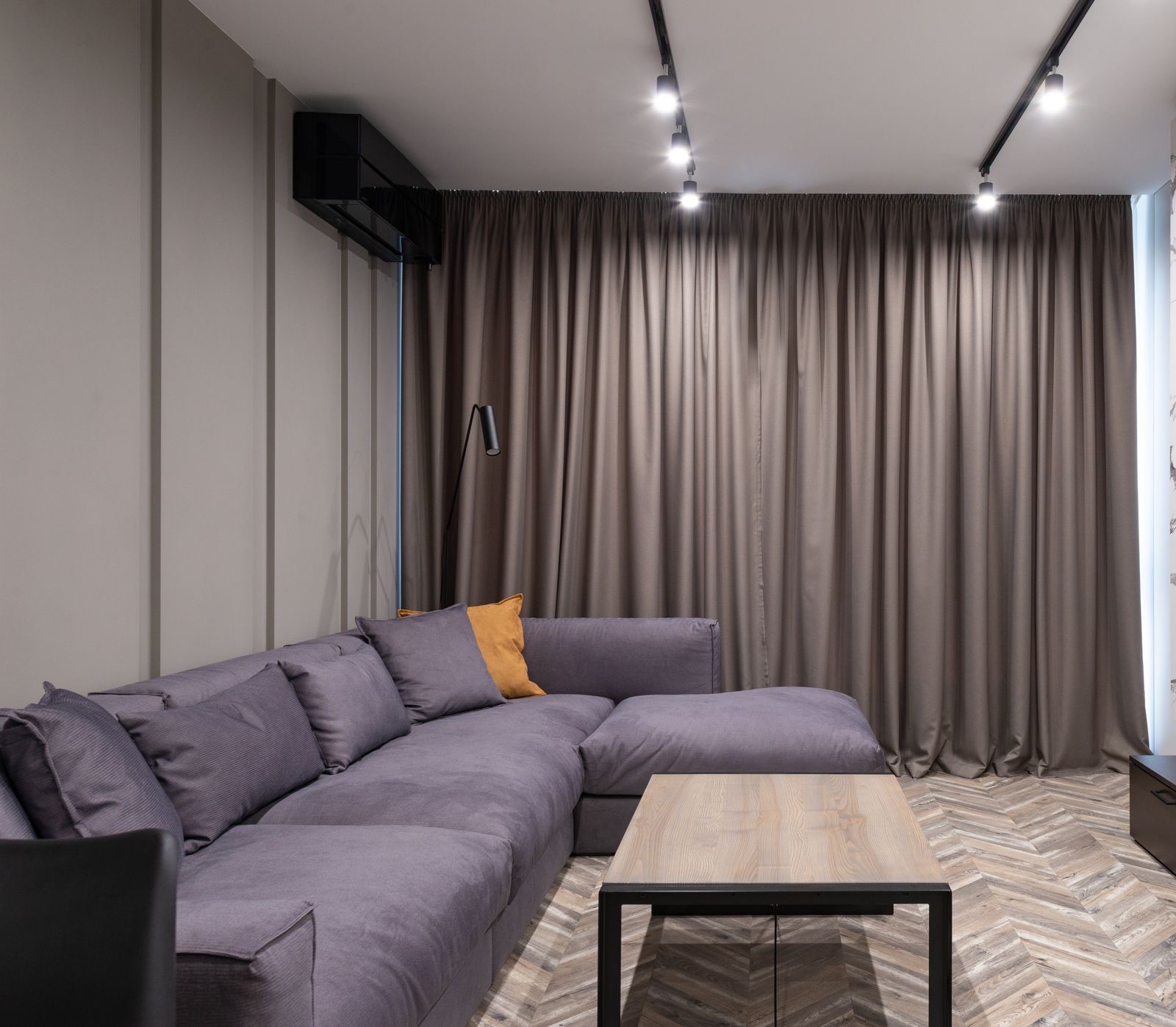 Living room with no false ceiling: two black surface-mounted track lights with adjustable spots run across a flat white ceiling, a black split AC sits high on the side wall, and floor-length taupe curtains hang from the ceiling line behind a grey corner sofa and a wooden coffee table on a herringbone floor.