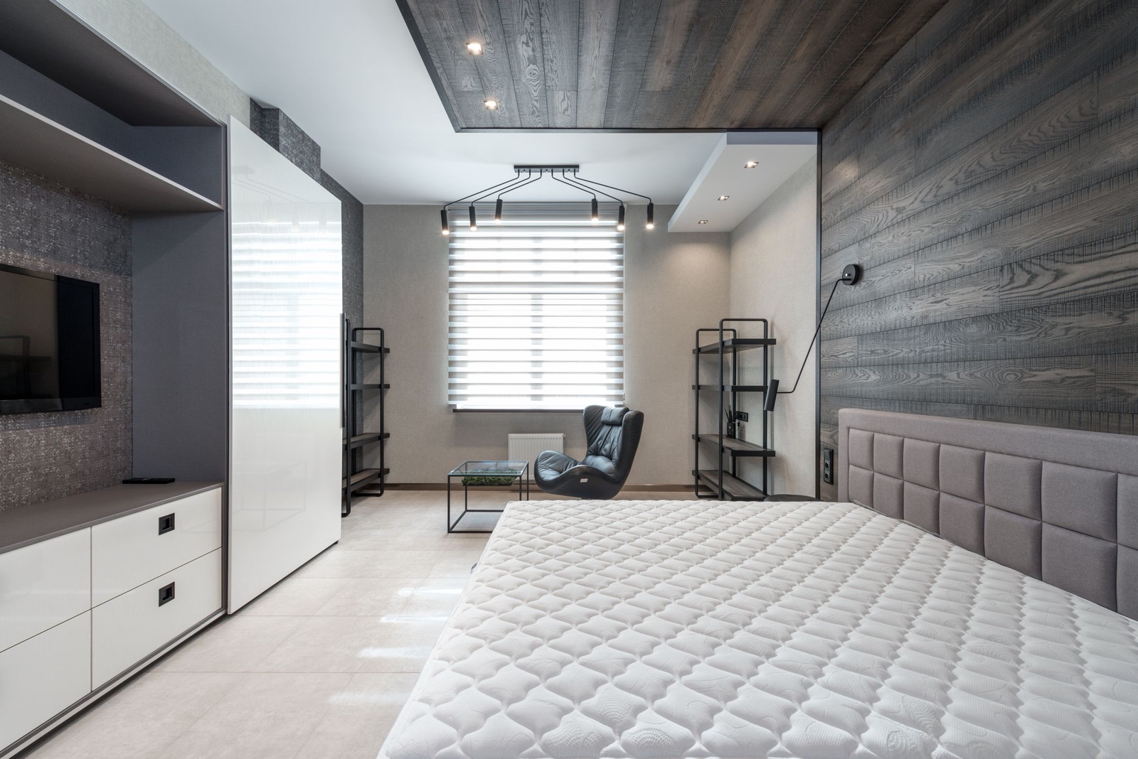 Grey and white bedroom with a TV mounted in an open bay of a built-in unit, white drawers below the bay, a tall white gloss sliding wardrobe next to it, a dark wood-panelled wall behind the bed and a reading chair by the window
