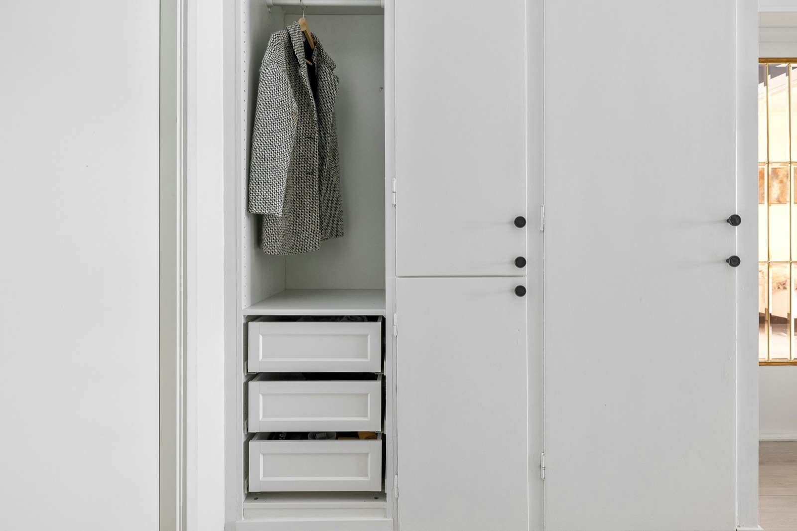 White fitted wardrobe with one bay open to show a hanging rail with a tweed coat above three white pull-out drawers, beside closed shutters with round black knobs