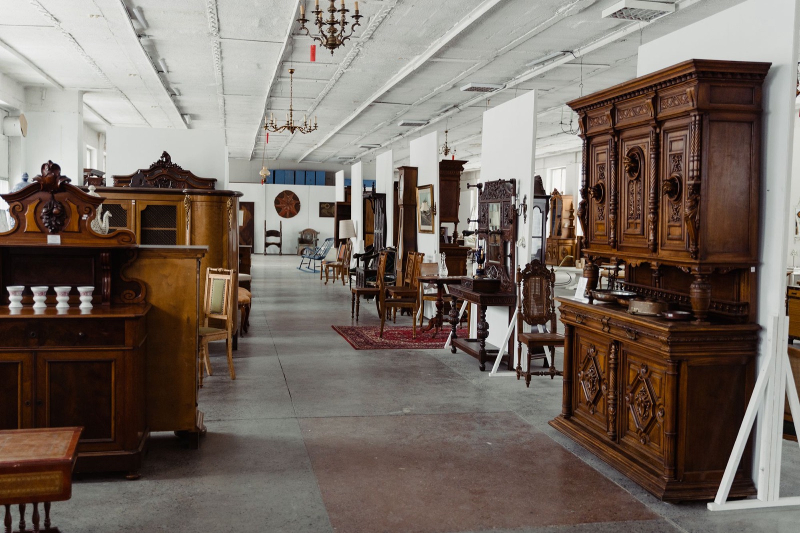 A long showroom aisle lined with carved dark-wood antique furniture: a tall sideboard with lion-head carvings, glass-fronted cabinets, cane-backed chairs, a grandfather clock and brass chandeliers on a white ceiling