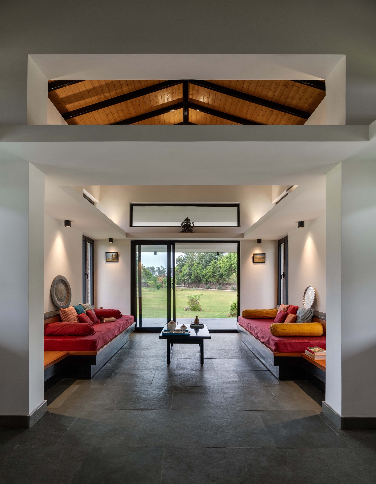 A long living room with two built-in diwans facing each other across a low black table, red mattresses with red and yellow bolsters, a dark natural stone floor, brass platters on the walls and a sliding door opening to a lawn, with a ventilator above it