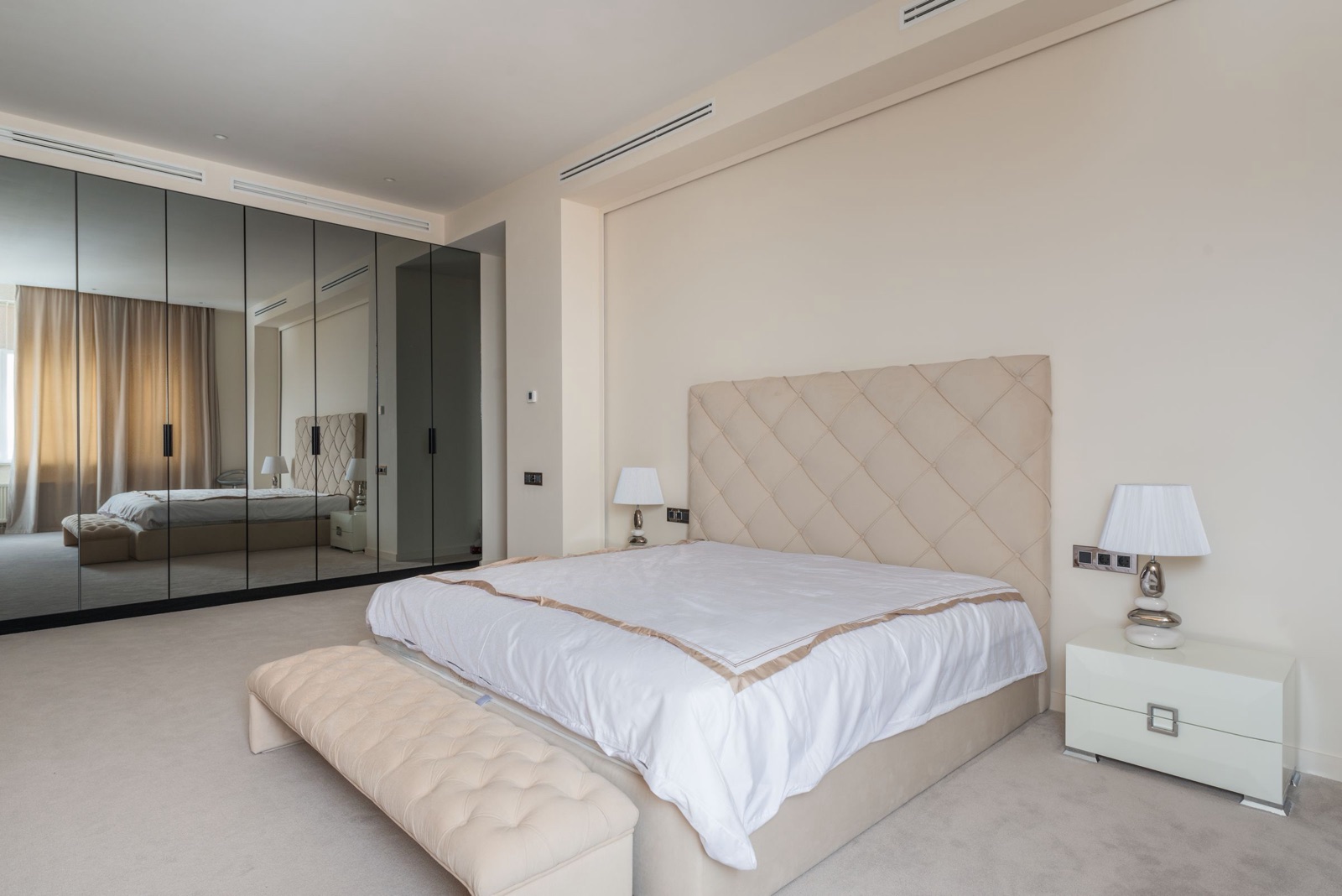 A large bedroom with a full wall of hinged wardrobe shutters faced in bronze-tinted mirror, slim black handles on each pair, reflecting the cream upholstered bed, bench and curtains opposite; a white bedside table and lamp stand beside the bed.