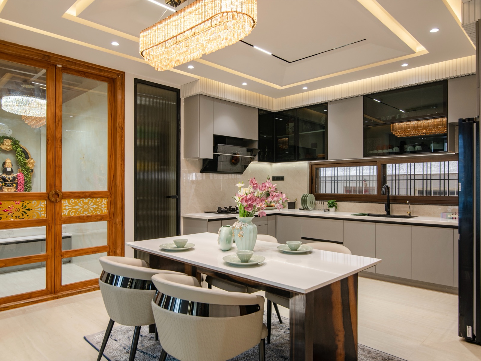 A four-seat rectangular dining table with a white top and dark wood base, set with pale green crockery and a vase of pink lilies, placed right beside an L-shaped kitchen with grey cabinets, a hob on one arm and a sink under a wide window on the other. To the left, carved wooden glass doors open to a lit pooja space.