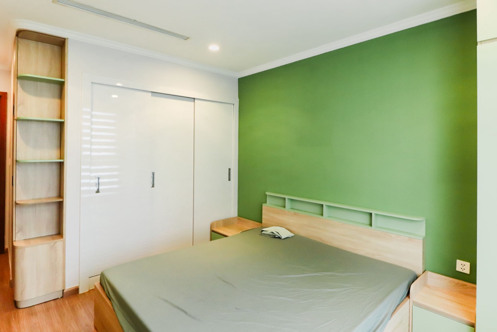 Bedroom with a green accent wall and a light oak laminate double bed whose headboard has a row of open mint-green cubbies, matching bedside units, a white gloss sliding wardrobe and a tall open shelf in the corner
