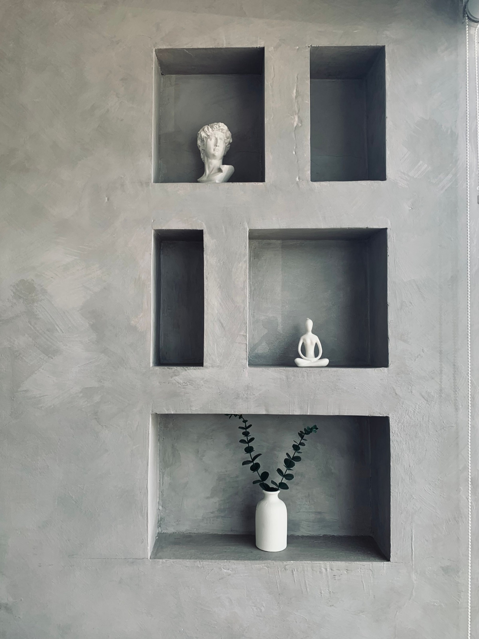 Five rectangular niches of different sizes recessed into a grey plaster wall, holding a white bust, a small white seated figurine and a white vase with eucalyptus stems