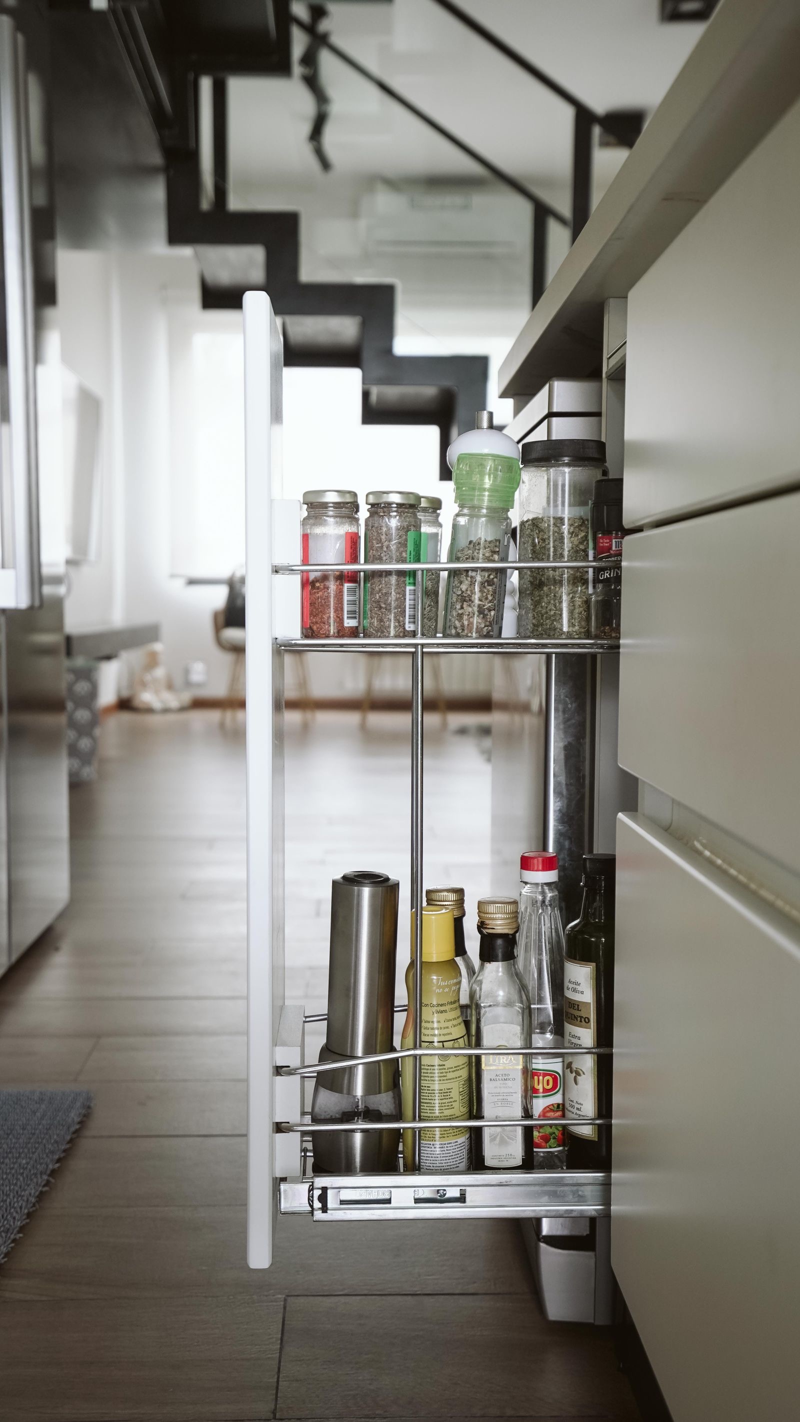 A narrow two-tier wire pull-out drawn out of a base cabinet, holding spice jars on the top tier and oil bottles on the bottom tier