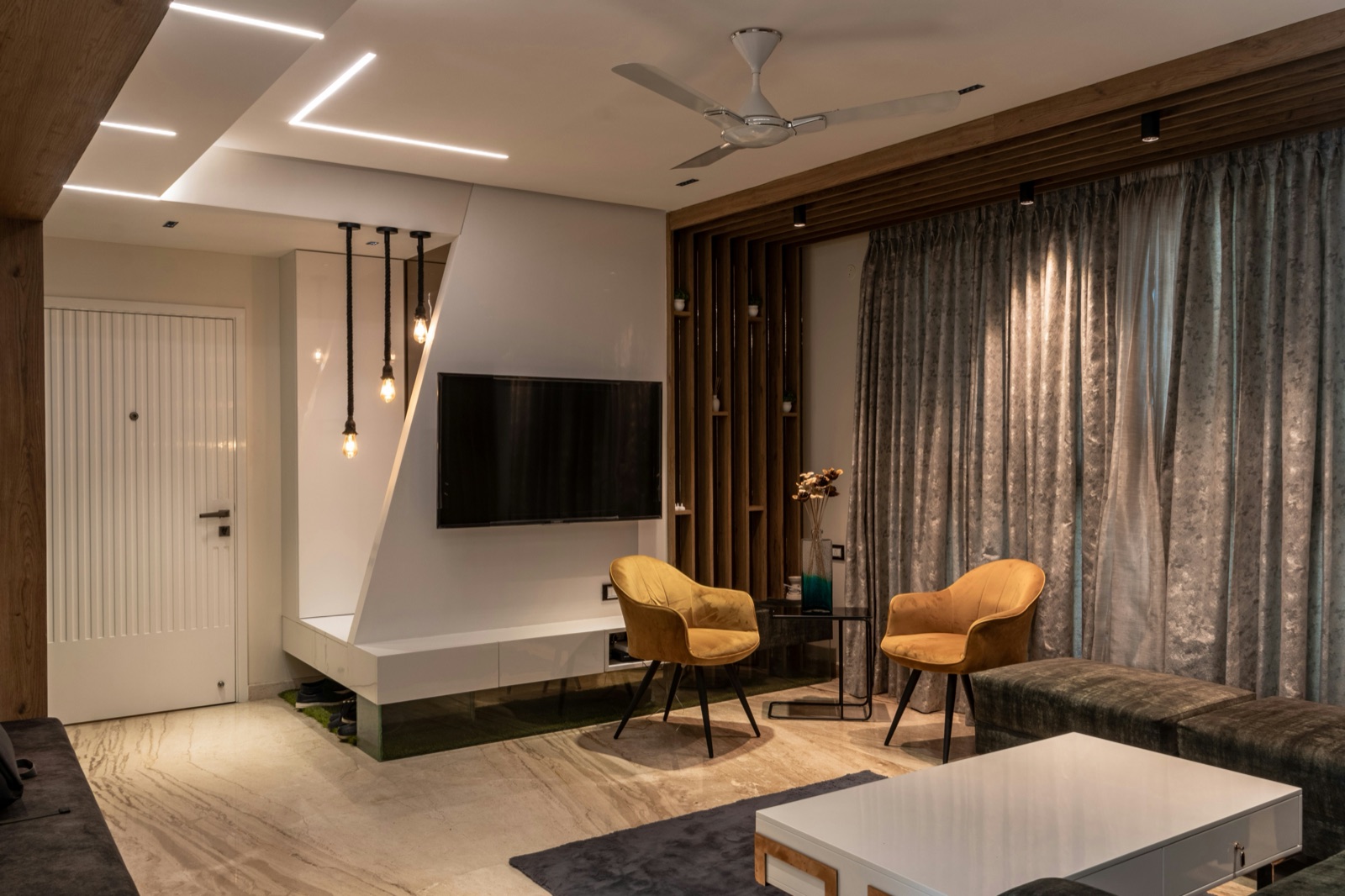Living room in a Chennai apartment with a marble-look stone floor, a white TV wall with a low floating console, a fluted white door, wooden vertical slats beside grey curtains, two mustard armchairs, linear profile lights set into the false ceiling and a white ceiling fan