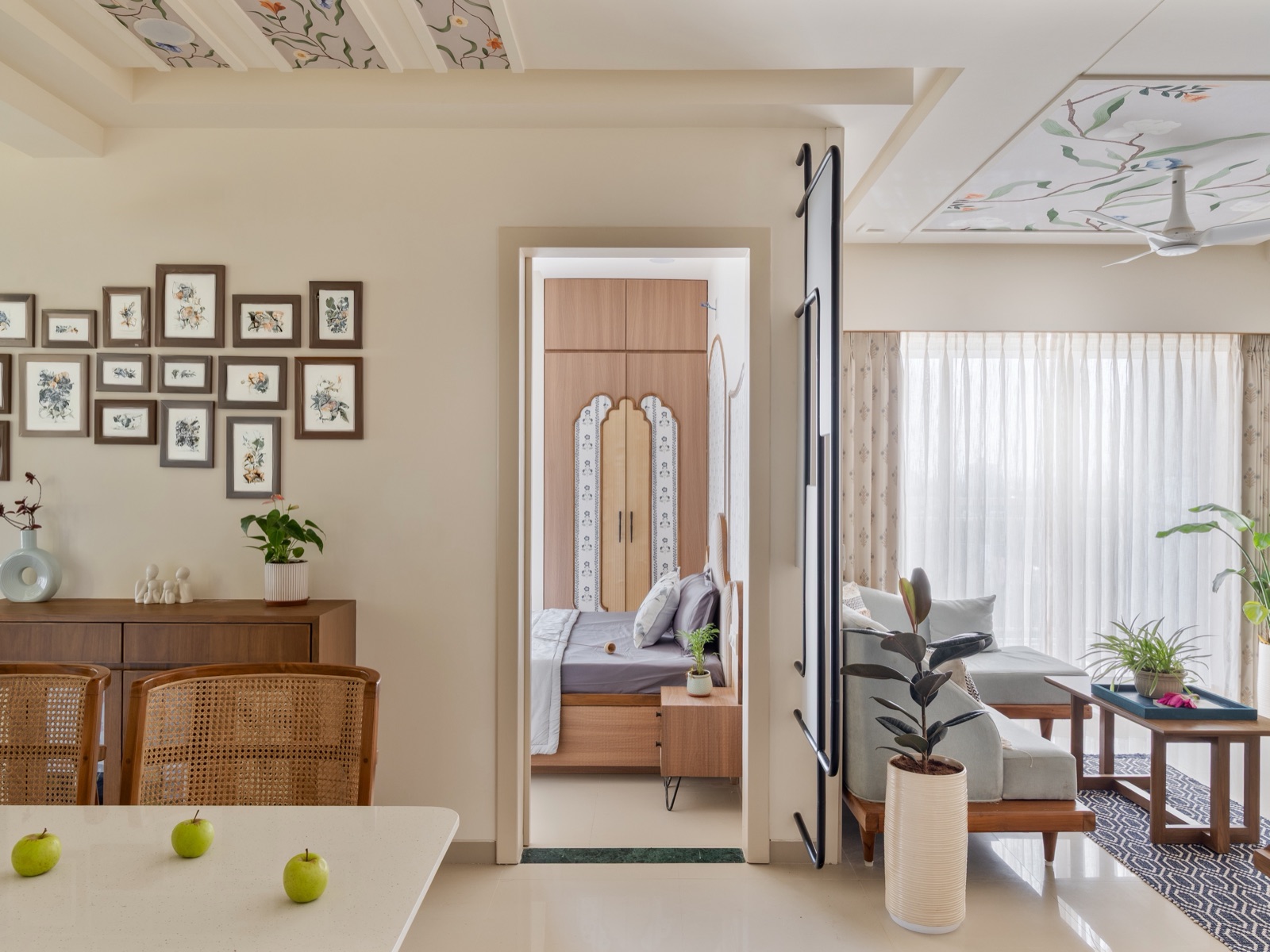 Apartment dining area with cane-backed chairs, a timber sideboard and a gallery wall of framed botanical prints, a doorway looking into a bedroom with an arched wardrobe, and a living area with a grey sofa and sheer curtains to the right, under painted floral ceiling panels