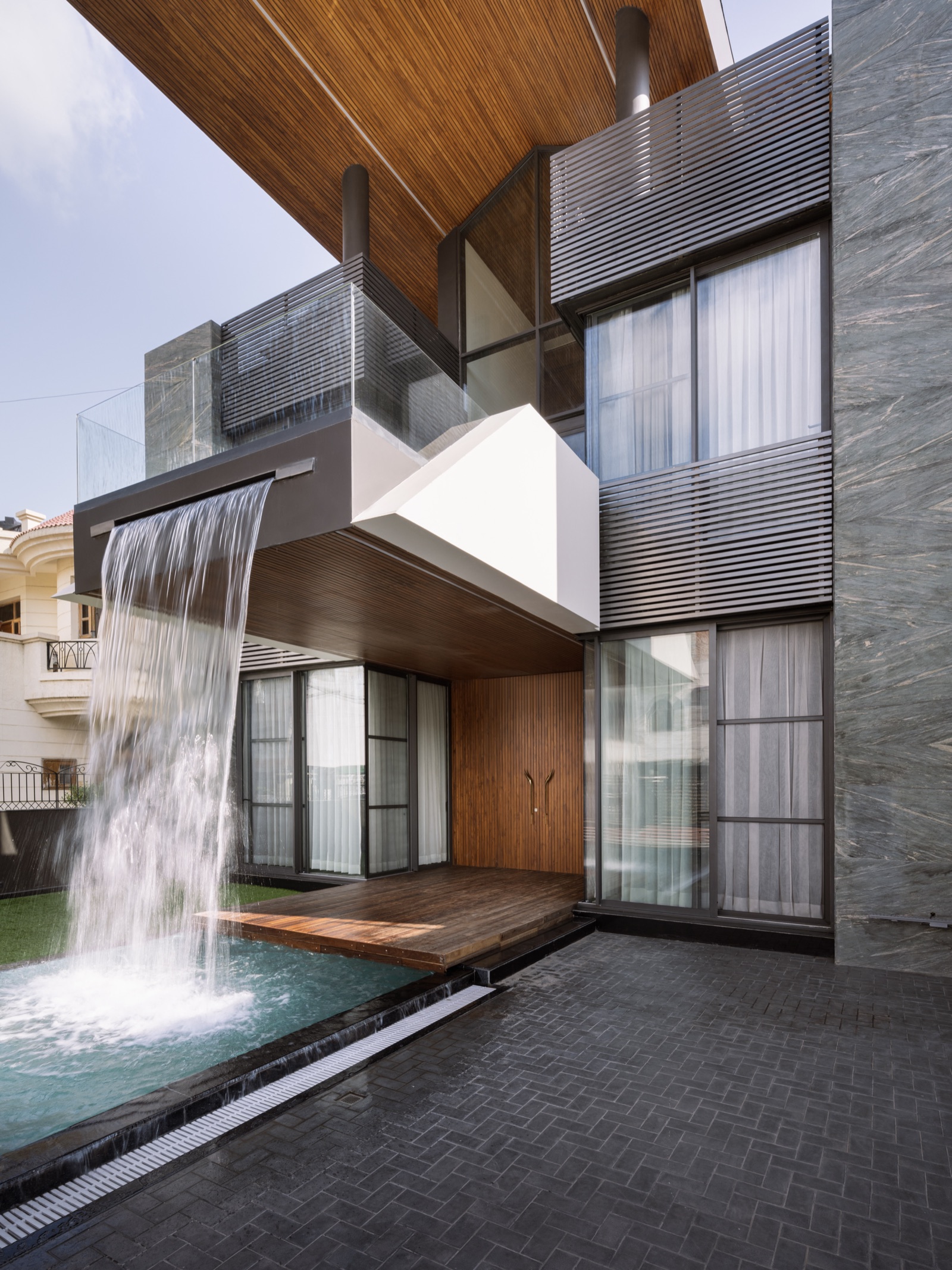 A cantilevered first-floor balcony with a frameless glass balustrade and charcoal horizontal louvres projects over a deck, with a sheet of water falling from its edge into a pool below and a timber-slatted soffit overhead
