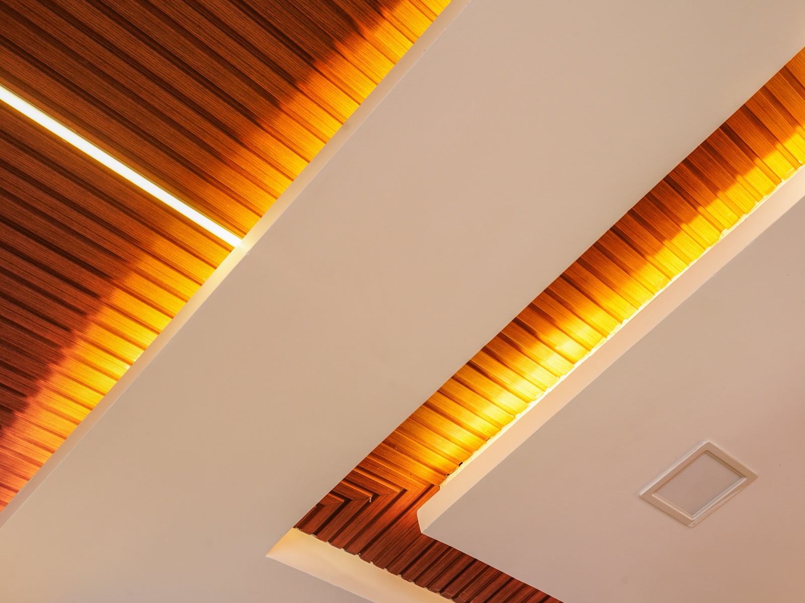 Close view looking up at a living room false ceiling where a band of narrow wooden slats meets a plain light-coloured gypsum panel, with a warm LED cove glowing along the joint and a small square ceiling grille.