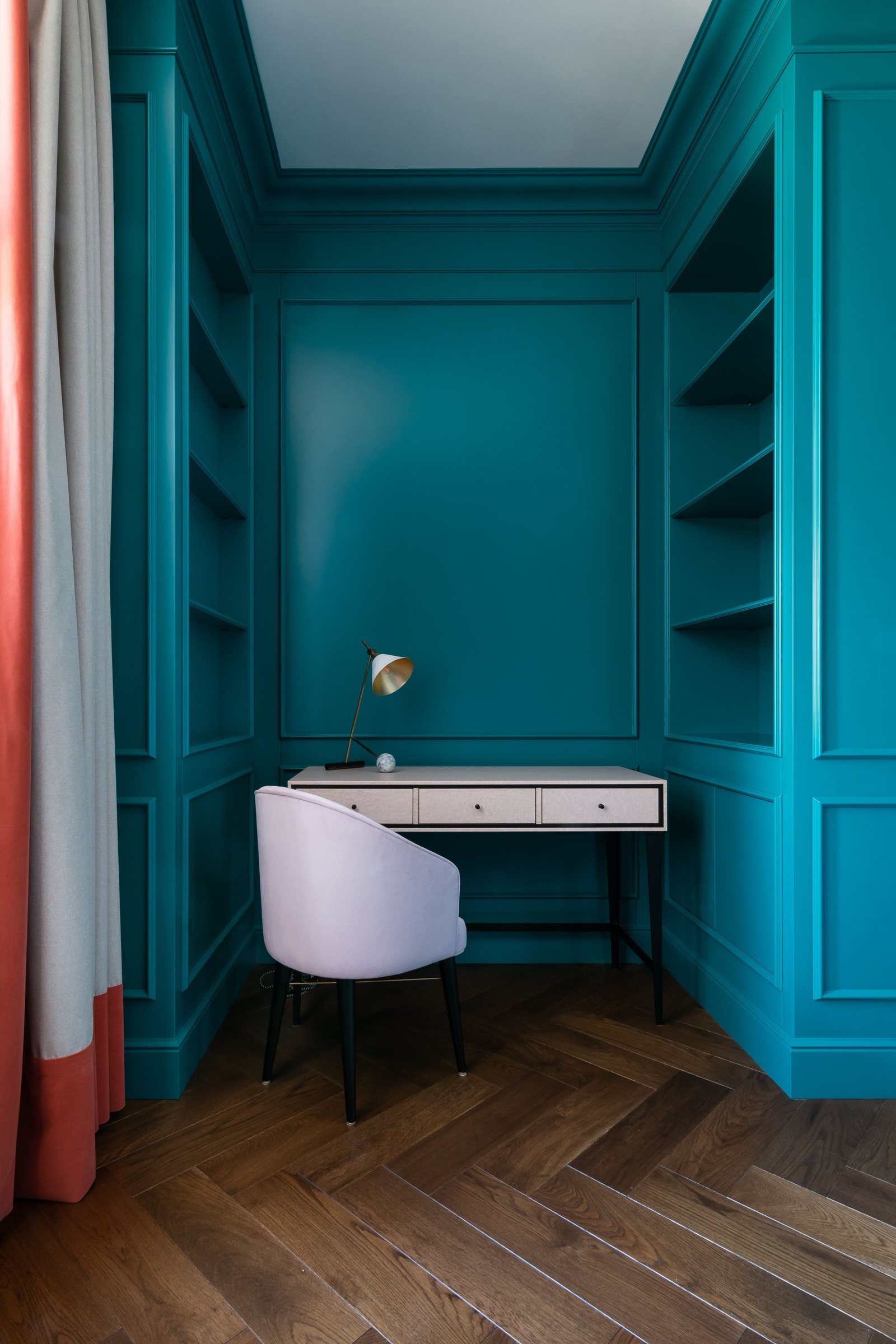 A study niche painted deep teal: a slim white desk with three small drawers and a brass task lamp set against a panelled back wall, with built-in open shelves on both sides, a lilac upholstered chair and a herringbone wood floor.