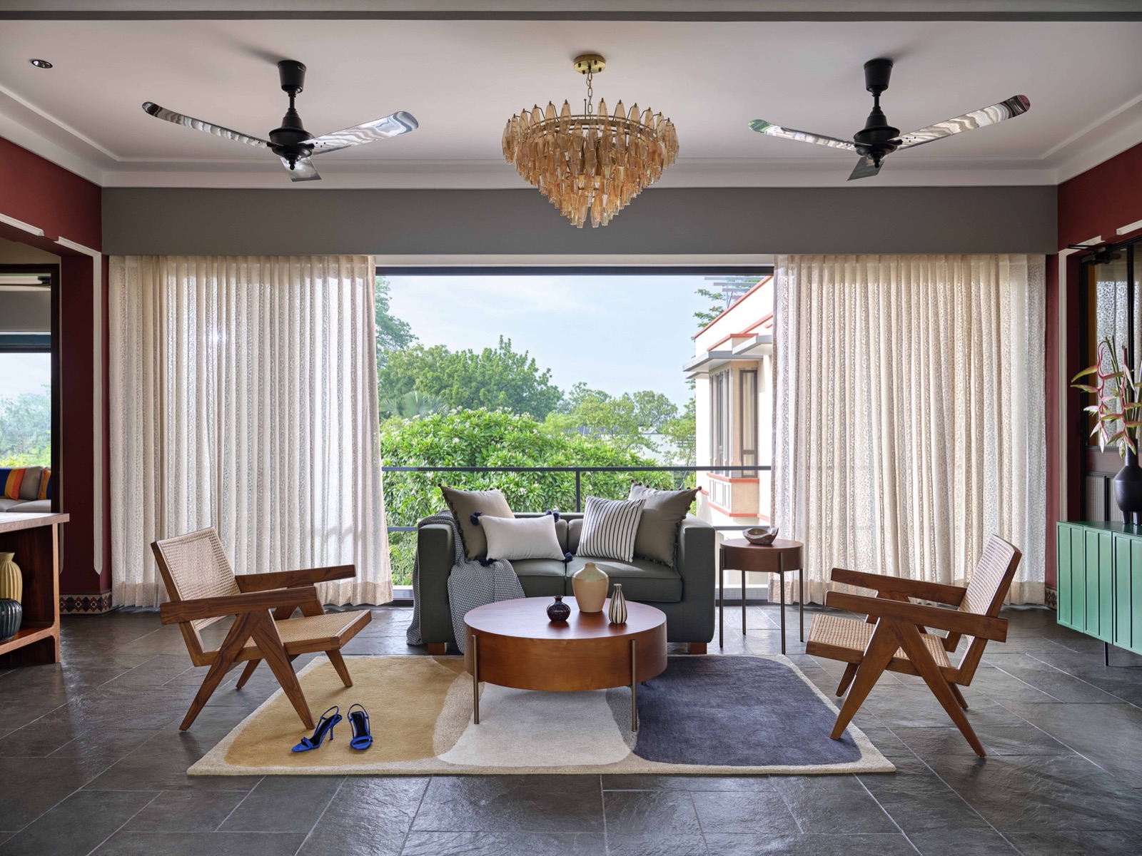 A living room with a grey two-seater sofa and two cane armchairs around a round wooden coffee table on a pale rug, slate floor, sheer curtains drawn back from a full-height opening onto a balcony and trees, maroon side walls and a crystal chandelier between two ceiling fans