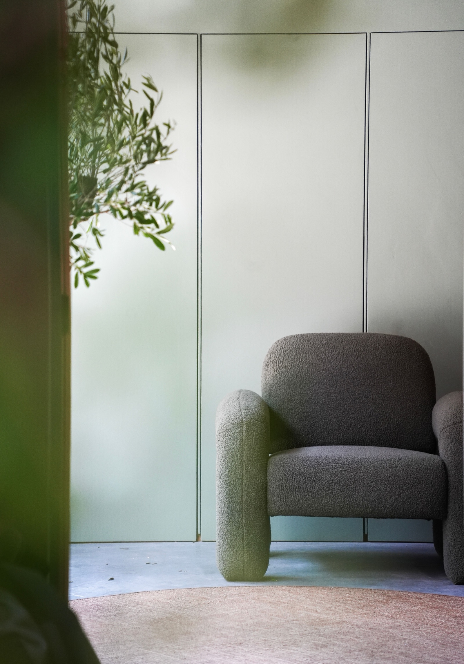 Pale sage green flush-panelled wall with a grey boucle armchair on a concrete floor, a soft pink round rug and an olive branch in the foreground