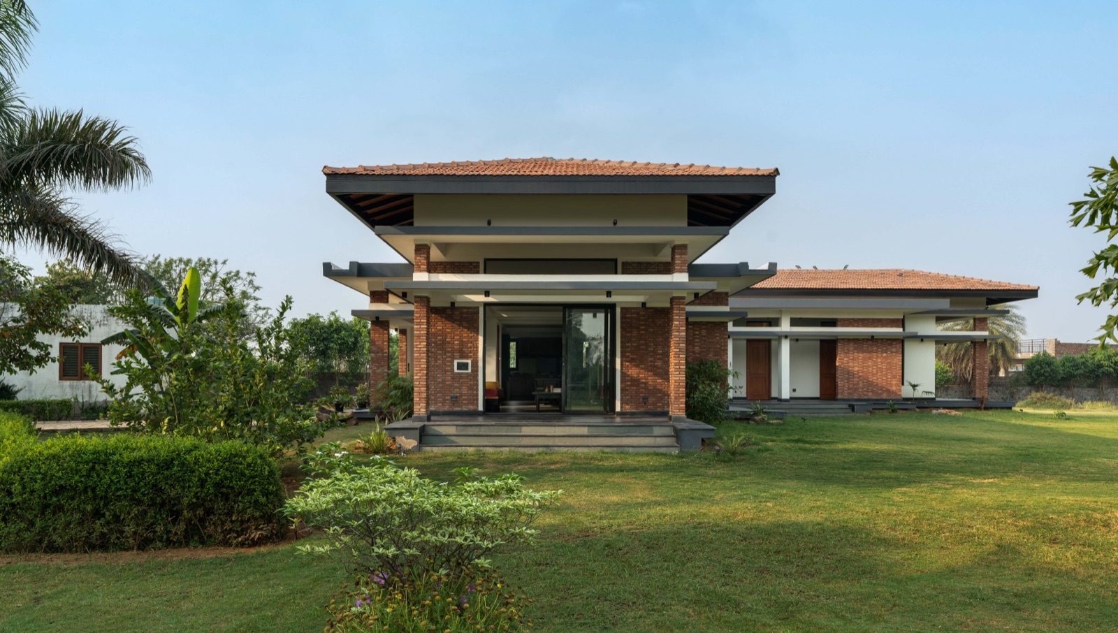 Single-storey house set in a lawn, with exposed red brick piers and walls, white bands, a central pavilion under a clay-tiled four-way pitched roof with deep overhangs, and a lower tiled wing to the right