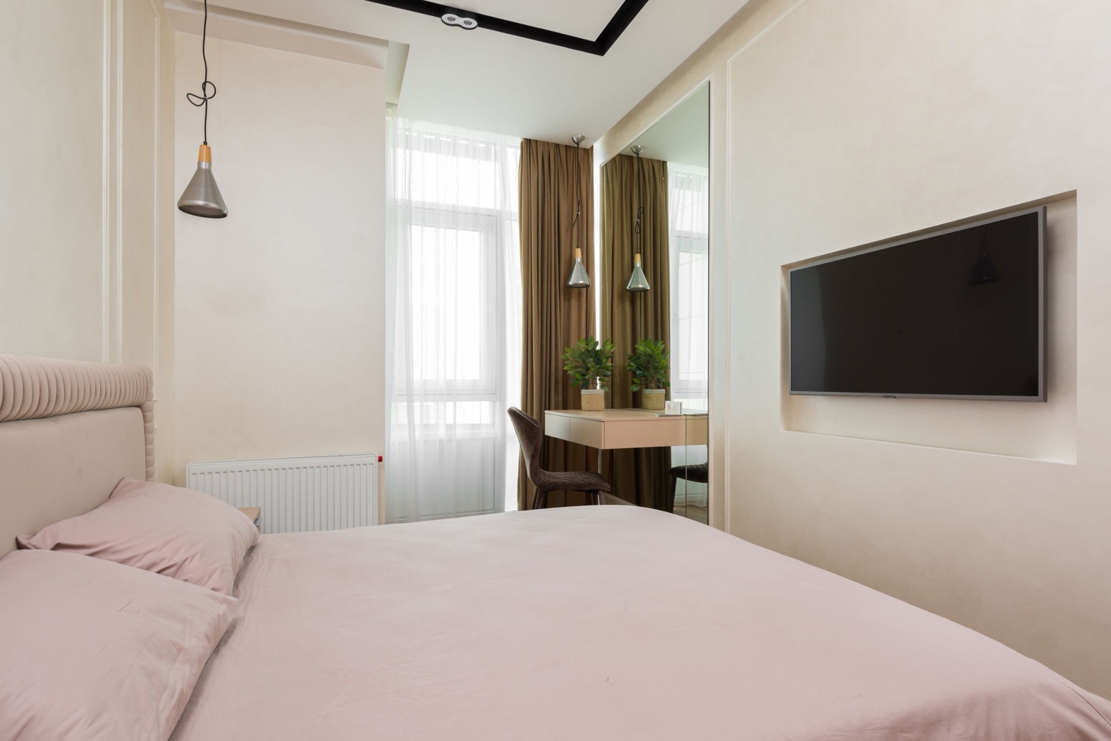 Bedroom with a pink bed in the foreground and a wall-mounted TV set into a shallow recess in a cream plastered wall, with no console below it; a slim dressing desk and a full-height mirror stand by the window beyond