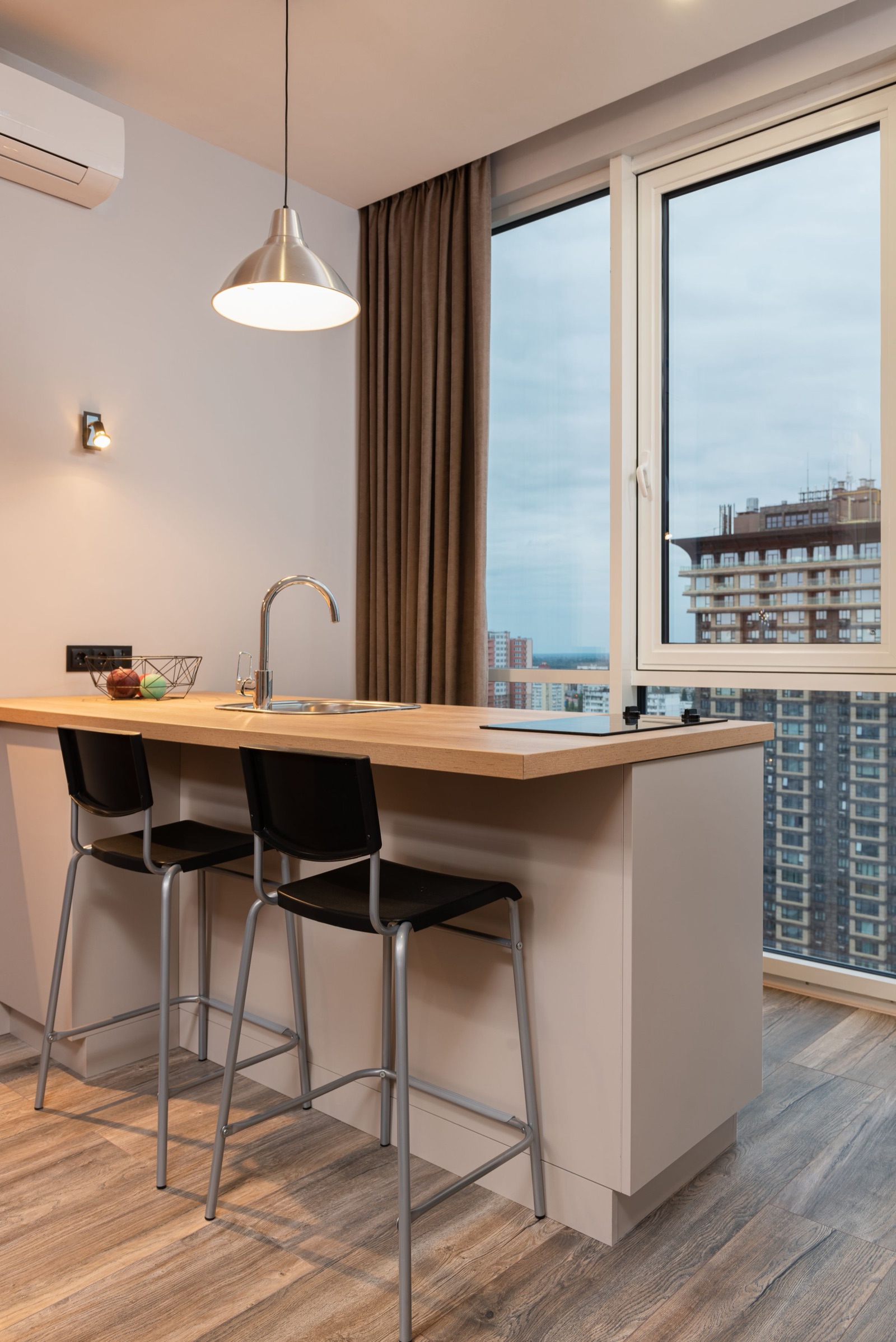 Small kitchen in a high-rise flat with a peninsula counter in wood-finish laminate holding a sink and a glass induction hob, two black bar stools on the room side, a metal pendant lamp above and a full-height window looking out at apartment towers.