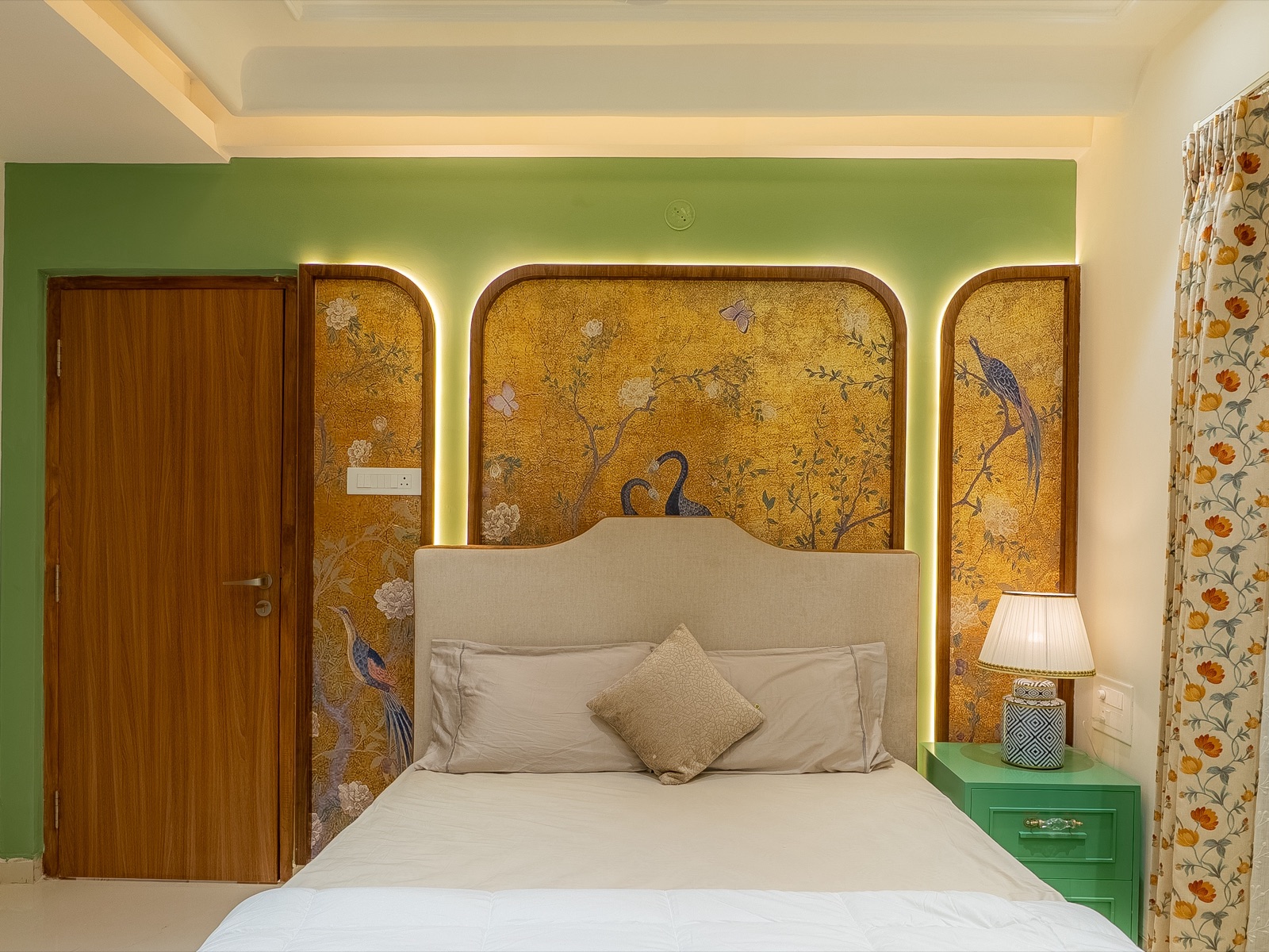 Bedroom with an upholstered headboard set against three round-topped, backlit panels painted with gold botanical scenes and peacocks, on a green wall, with a green bedside table and floral curtains