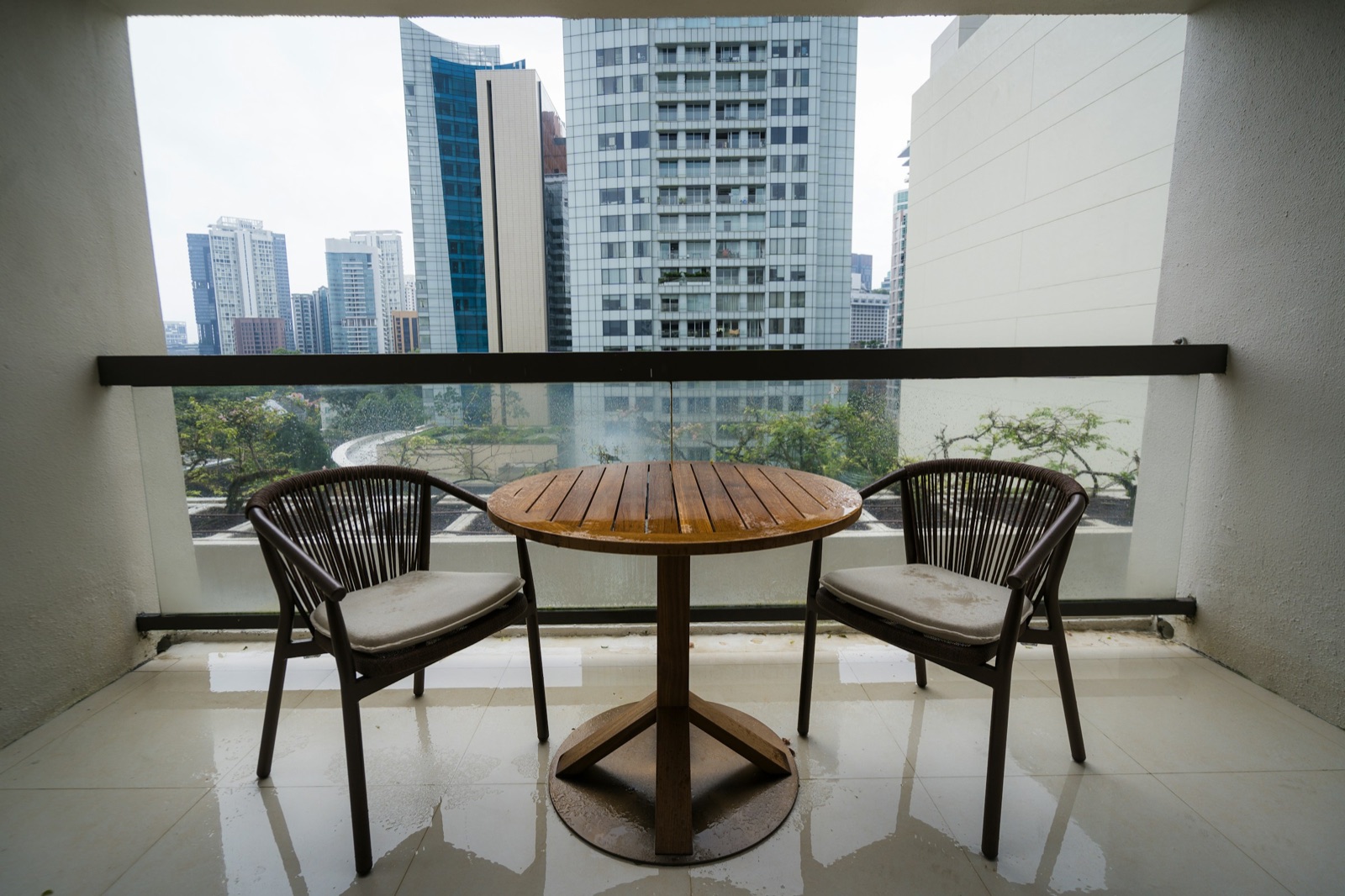 A narrow high-rise balcony with a pale tiled floor, a solid parapet topped by a glass panel and a dark handrail, and a small round wooden bistro table between two woven outdoor chairs with cushions, looking out over city towers