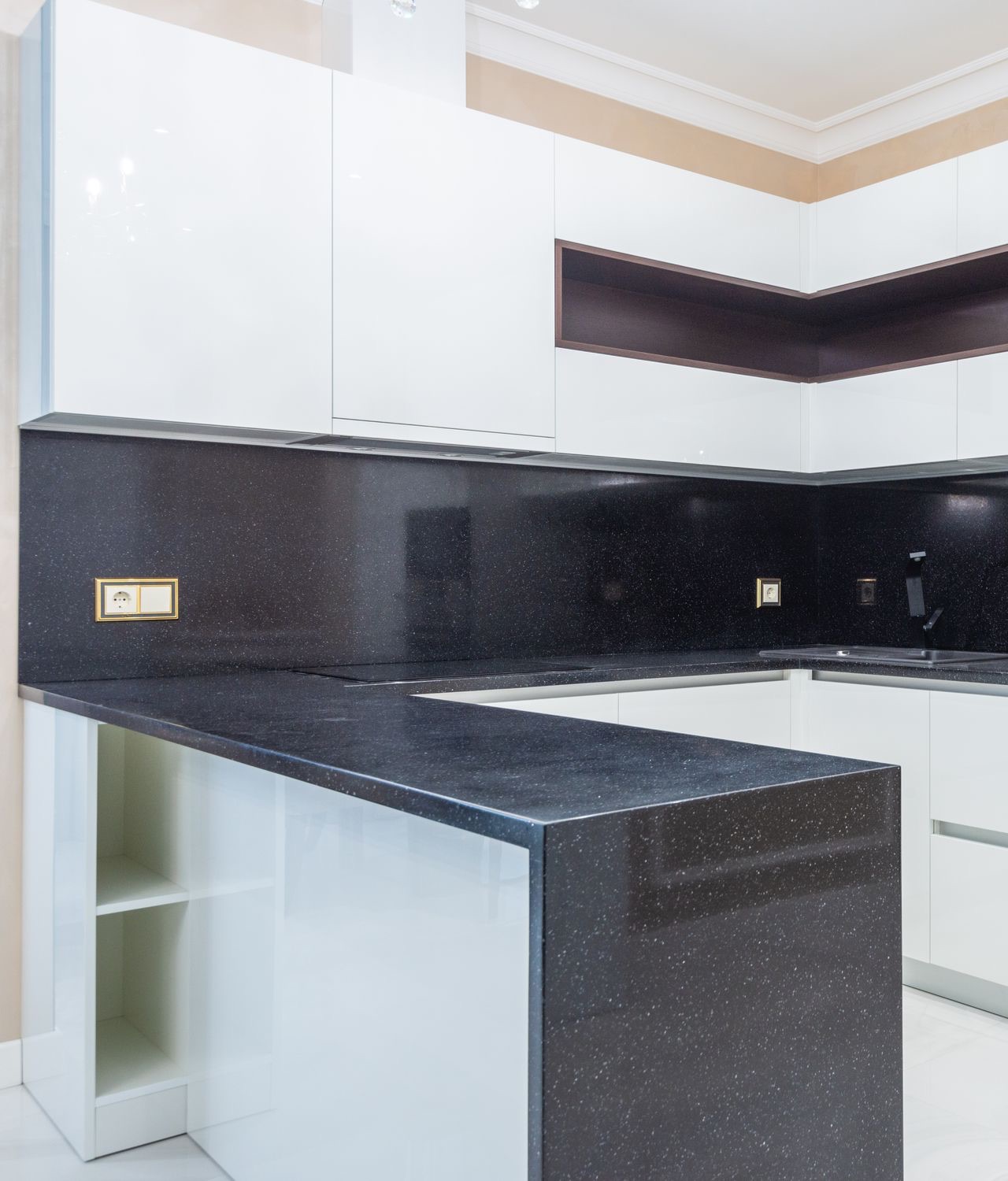 L-shaped kitchen with a black speckled stone platform that turns down to the floor at the end of a peninsula, a full-height splashback in the same black material up to glossy white wall cabinets, and a flush hob and sink in the counter.