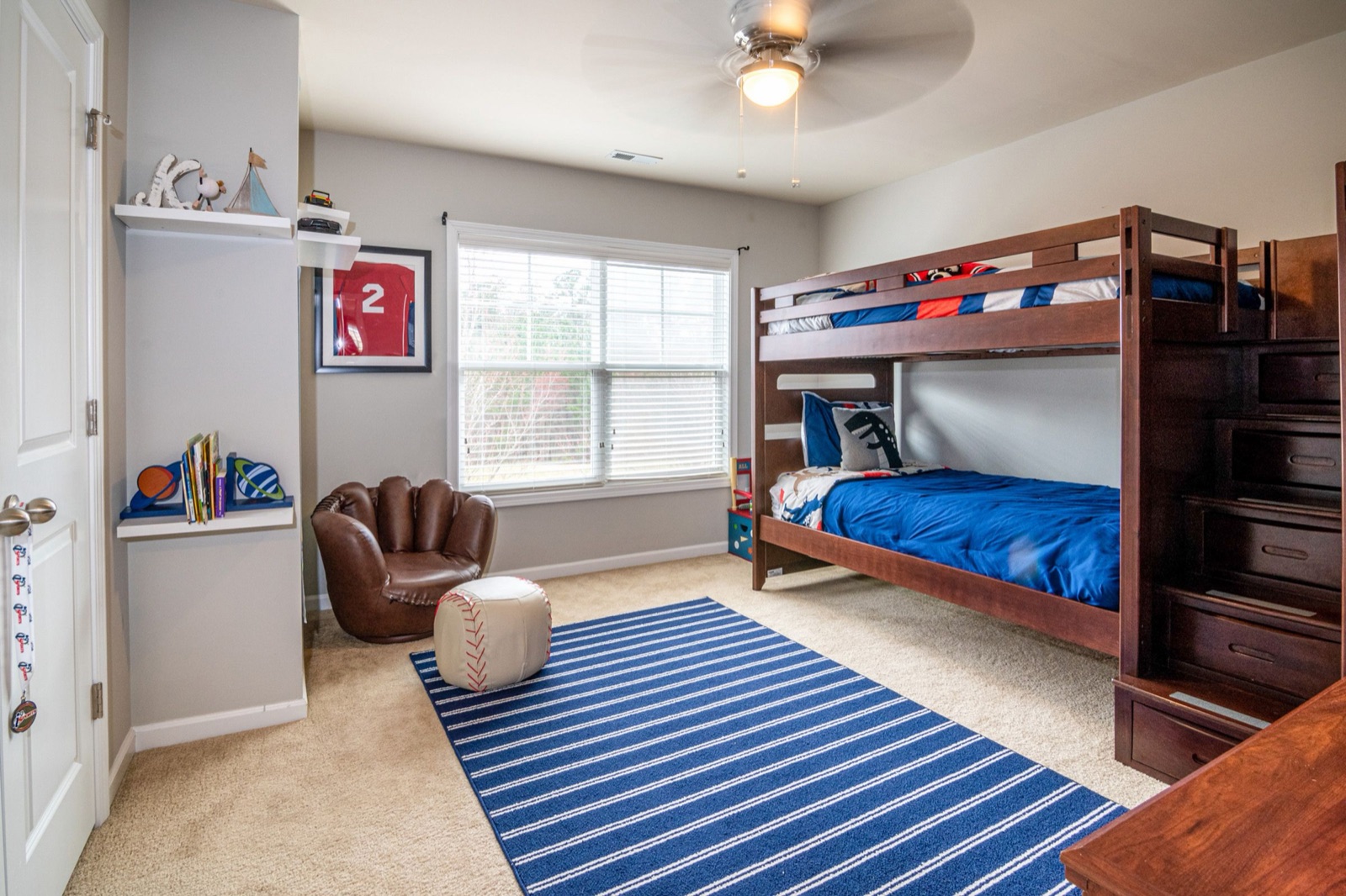 A kids bedroom with a dark wood bunk bed whose side is a staircase of deep drawers instead of a ladder, both bunks dressed in blue bedding, a striped blue rug, floating wall shelves holding books and a ceiling fan with a light.