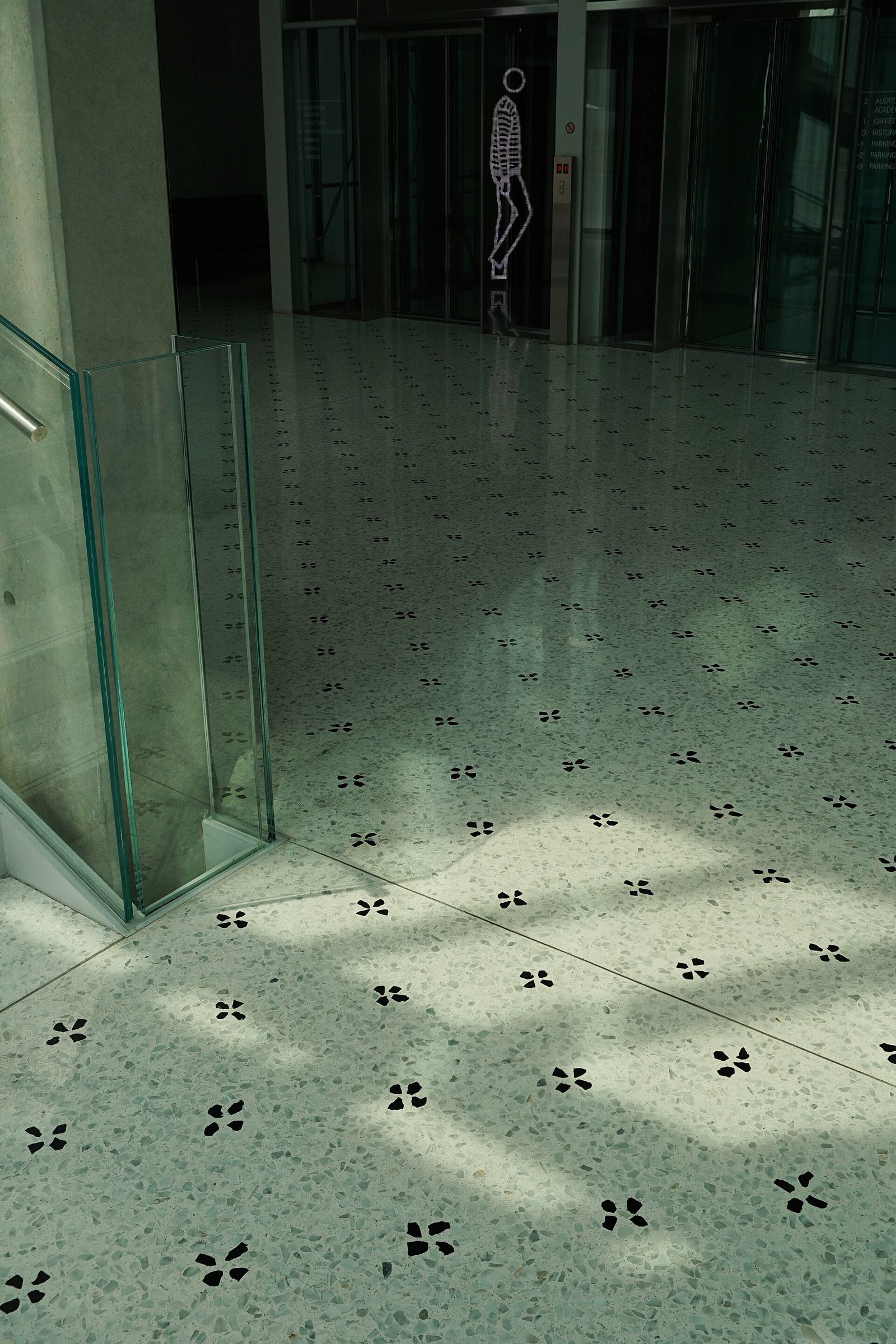 A pale terrazzo floor with grey and white stone chips and a repeating inlay of small black four-petal flowers, running from a glass-walled stair past a concrete column towards lift doors