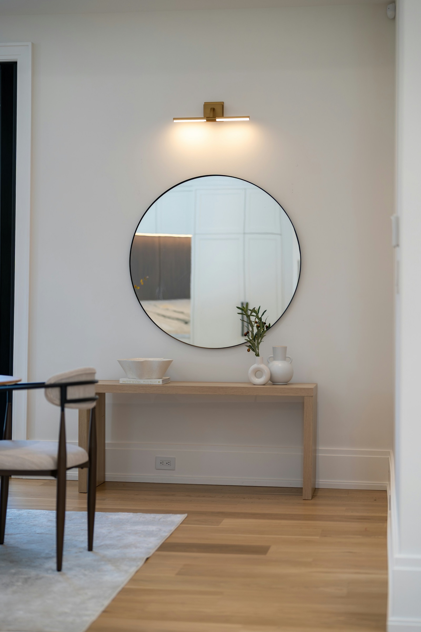 A large round mirror with a thin black frame hung above a pale oak console table holding a ceramic bowl, books, white vases and an olive sprig, with a brass picture light above the mirror, on a white wall by a wooden floor; a dining chair stands at the left.