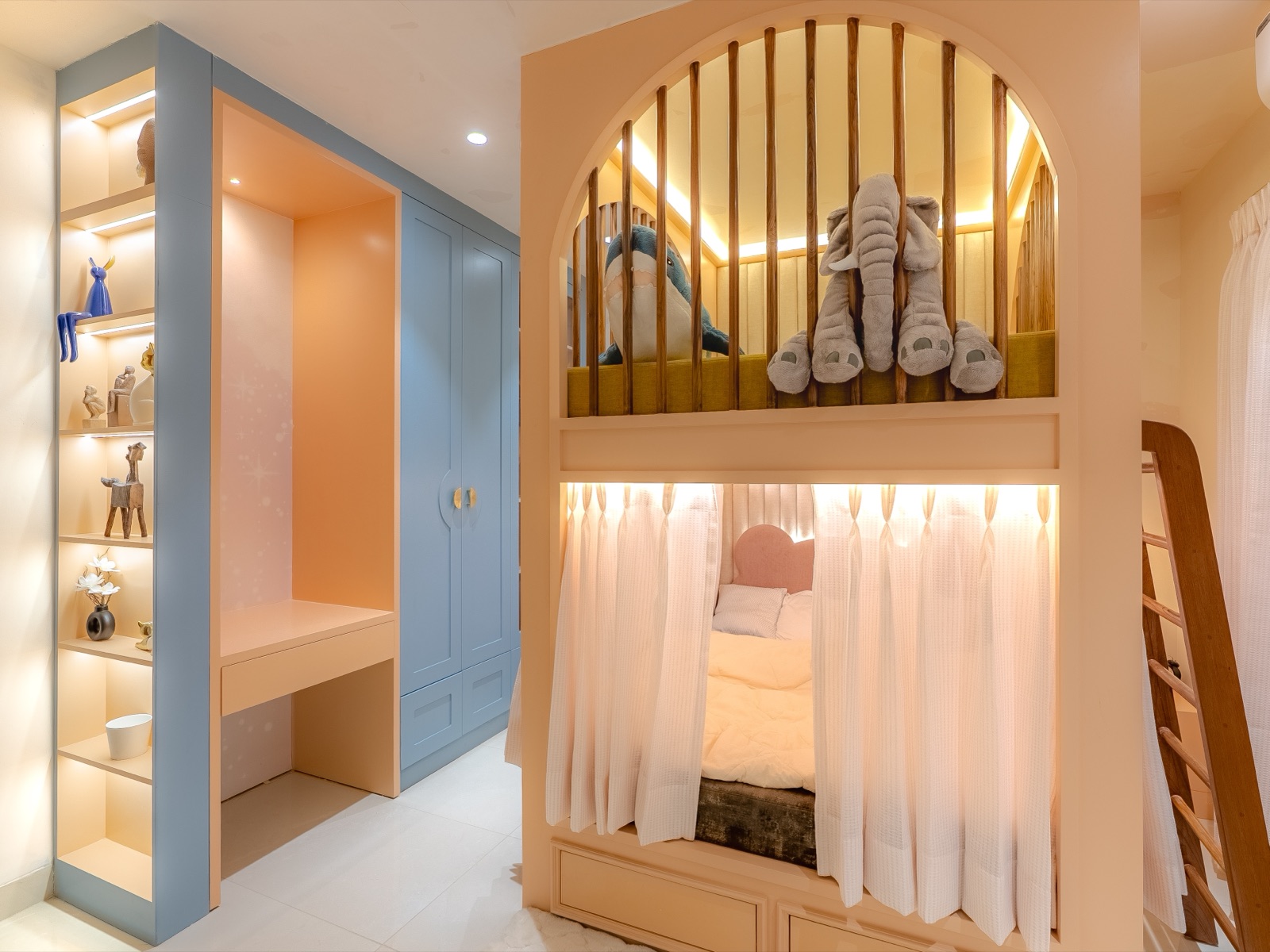 A pastel children's room with a built-in bunk unit in peach, the upper bunk behind arched wooden slats with two soft toys and a wooden ladder at the side, the lower bed screened by sheer curtains, and to the left a study nook with a desk and drawer, a powder-blue wardrobe with drawers below, and lit open display shelves.