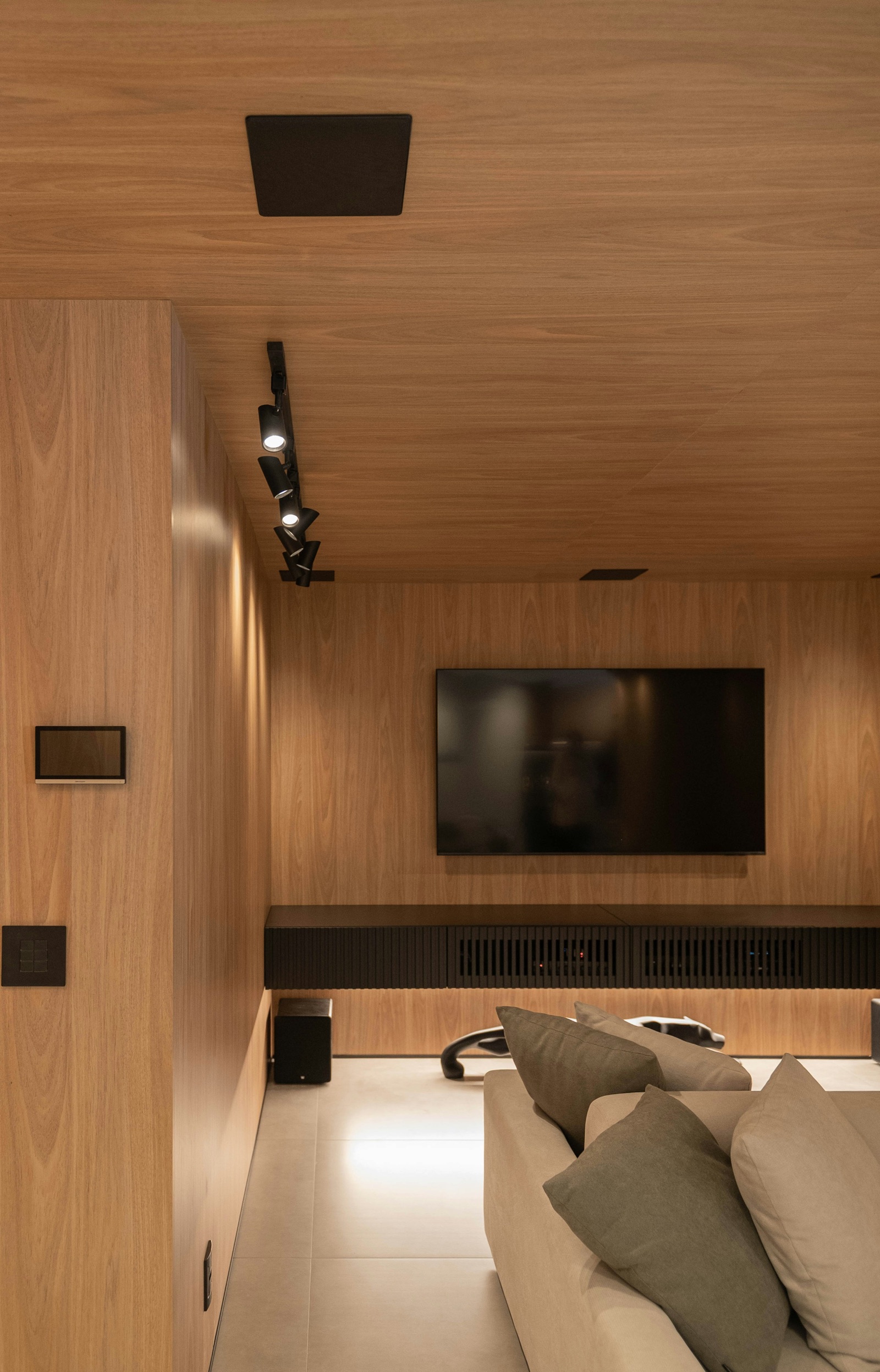 Photograph of a media room lined on walls and ceiling with matching light wood-grain panels, a wall-mounted TV above a long black slatted console, a black track light and a beige sofa in the foreground.