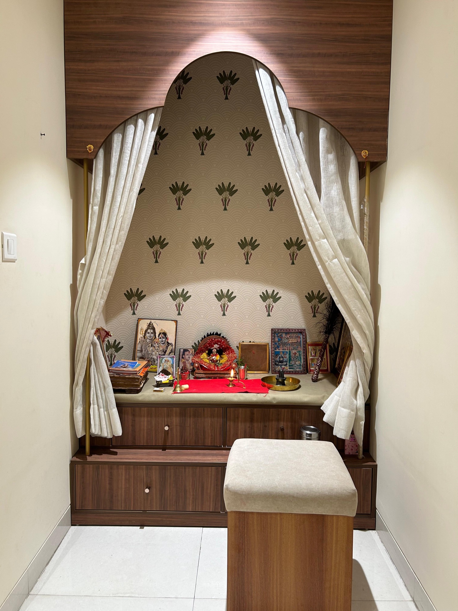 A pooja alcove at the end of a short passage in a Raipur flat: a walnut-laminate surround with a three-lobed arched opening, a wallpaper of small palm motifs inside, white linen curtains tied back on slim brass rods, a ledge with framed pictures, a brass lamp and a brass plate, two rows of drawers below and an upholstered stool in front.