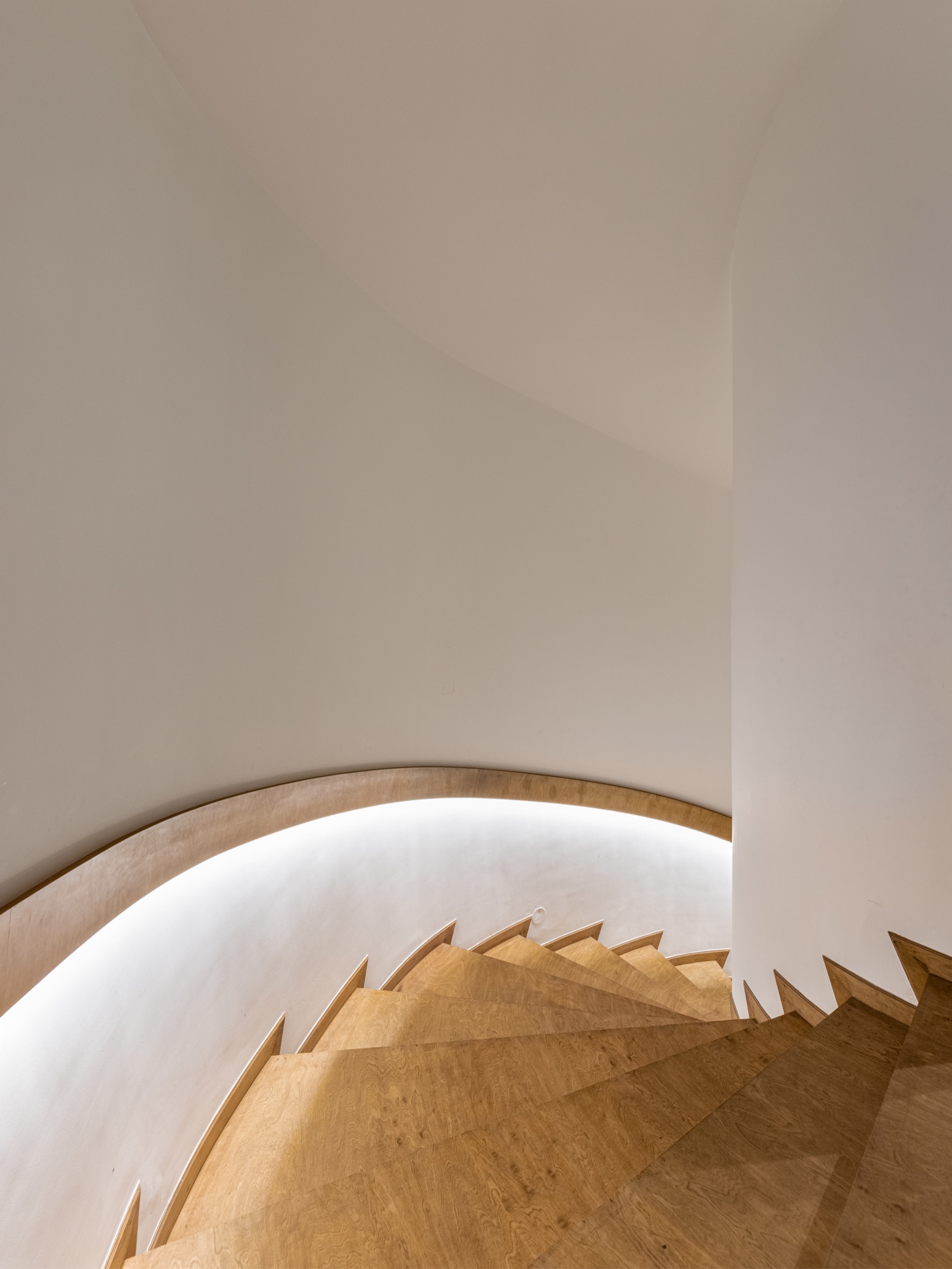 Looking down a curved staircase with light wood treads between smooth white curved walls, with a timber handrail set into a recessed groove in the wall and an LED strip lighting the steps from inside the groove