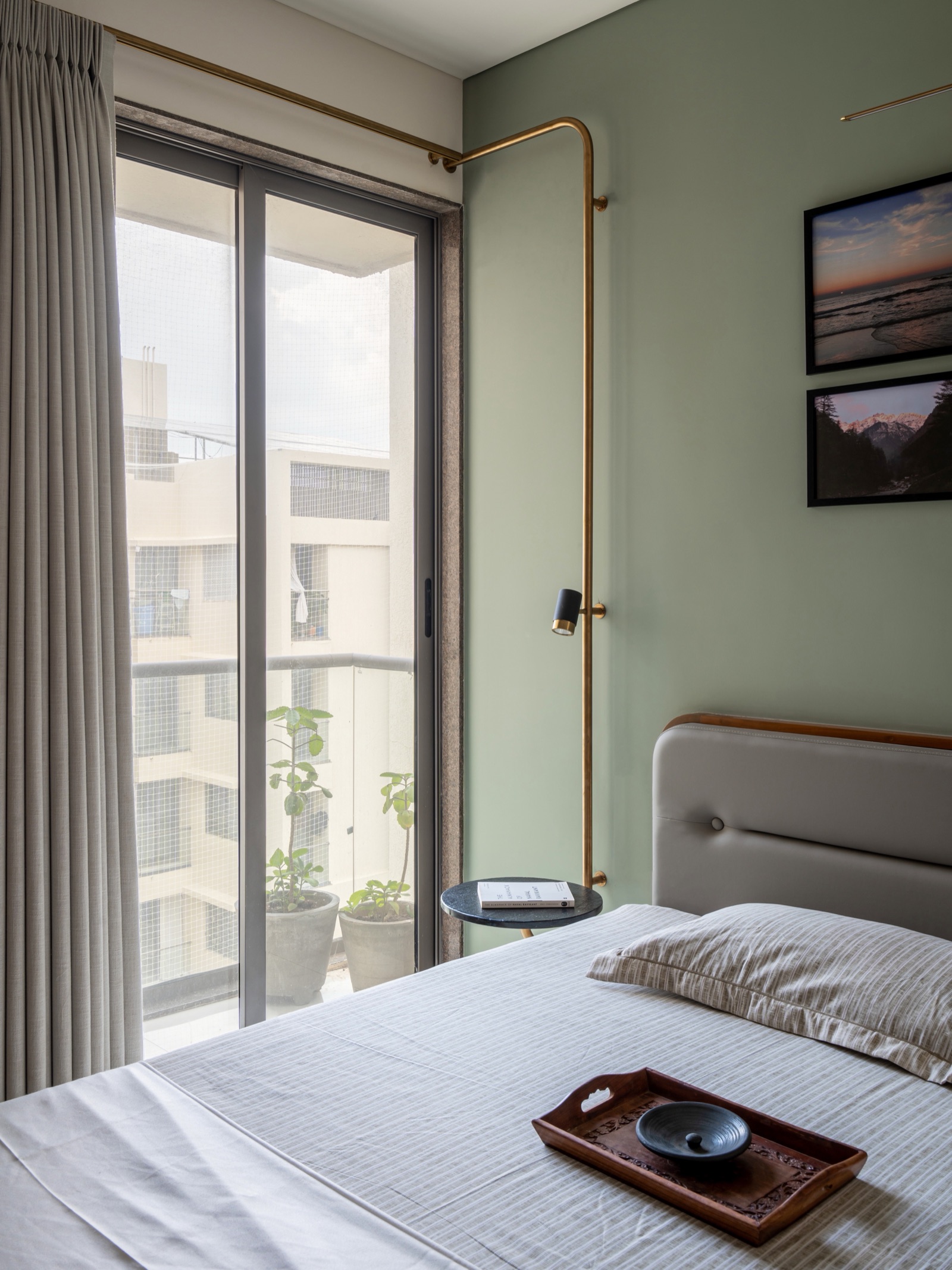 Bedroom with a pale sage green wall behind a grey upholstered bed, white ceiling, grey-beige full-height curtains beside a balcony door, and a slim brass pipe reading lamp