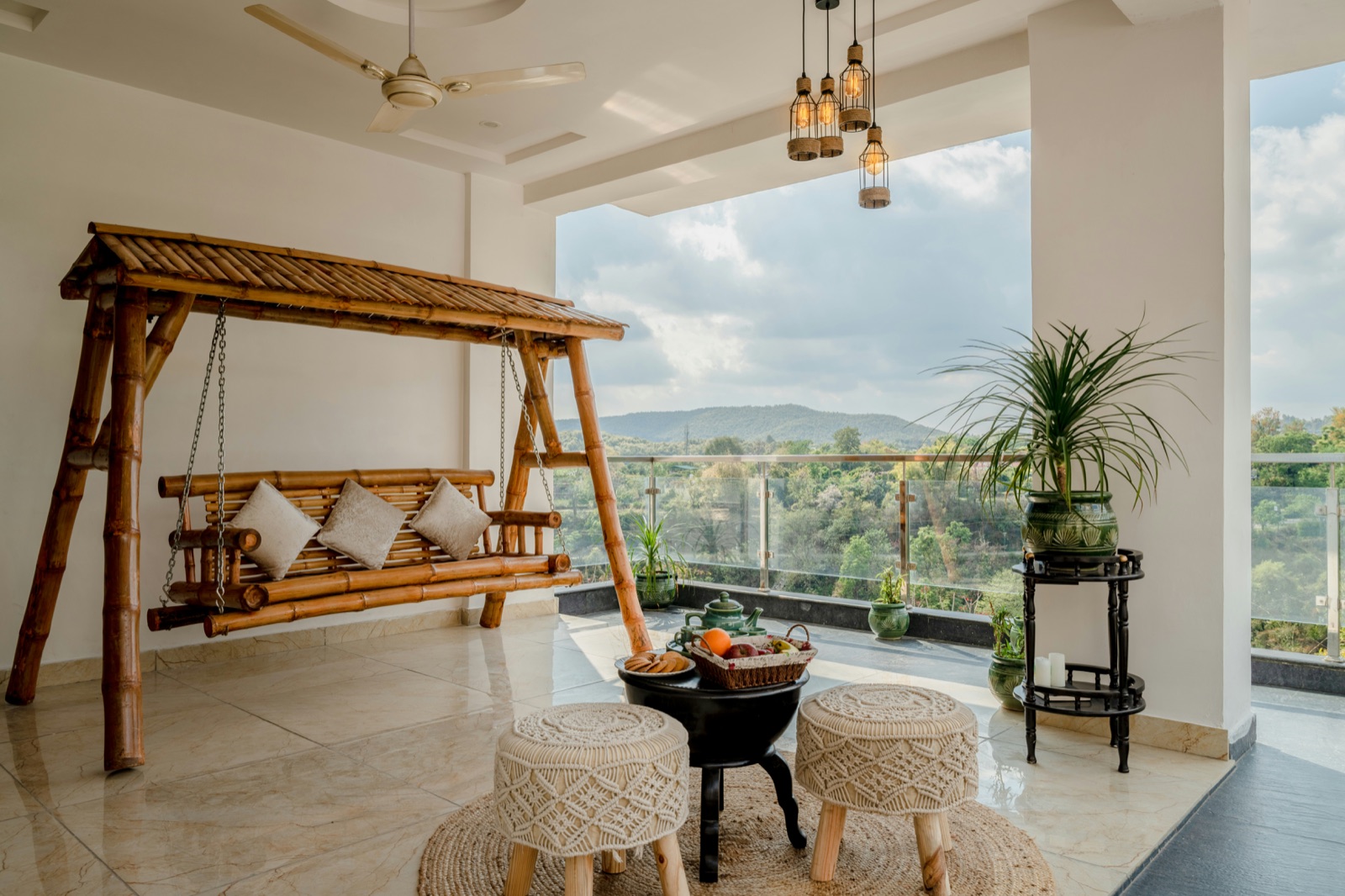 A wide covered balcony with a bamboo swing seat hung on chains from a bamboo frame with a slatted roof, cushions on the swing, two macrame-covered stools and a small black table on a jute rug, potted plants, a glass railing and wooded hills beyond