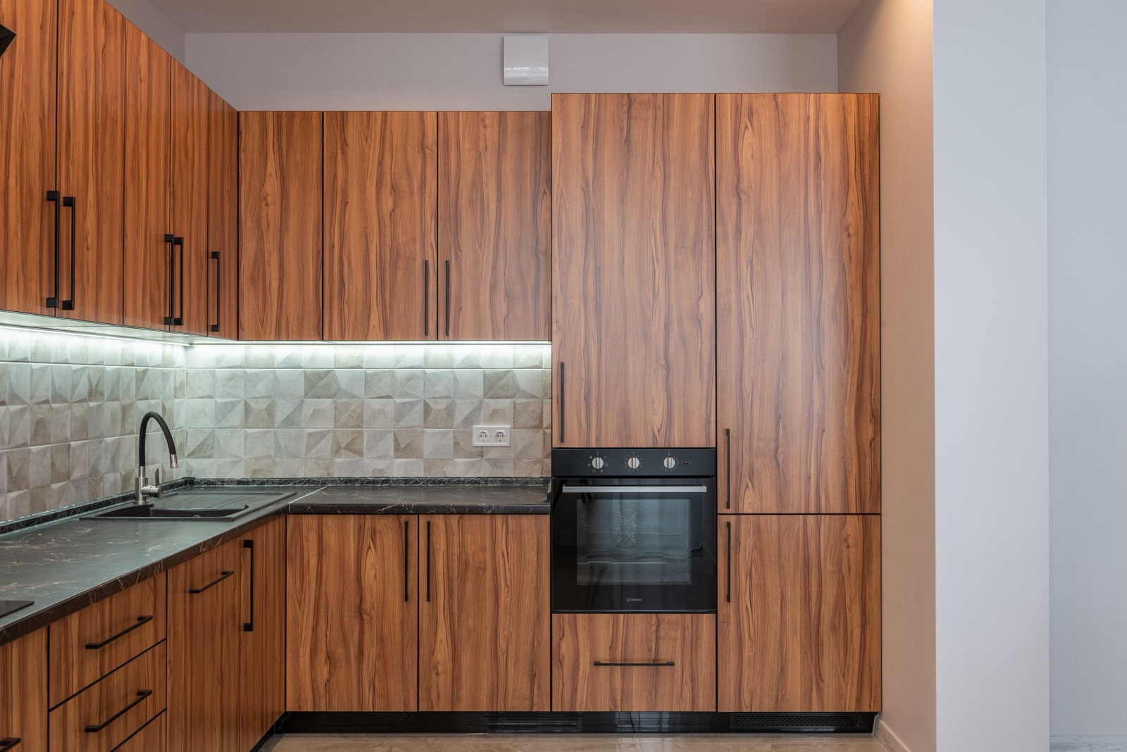 L-shaped kitchen in walnut-finish laminate with a black built-in oven set at counter height in a tall unit, a full-height two-door tall unit beside it, a ventilation grille in the plinth below, and a double socket on the tiled wall above a dark stone counter.