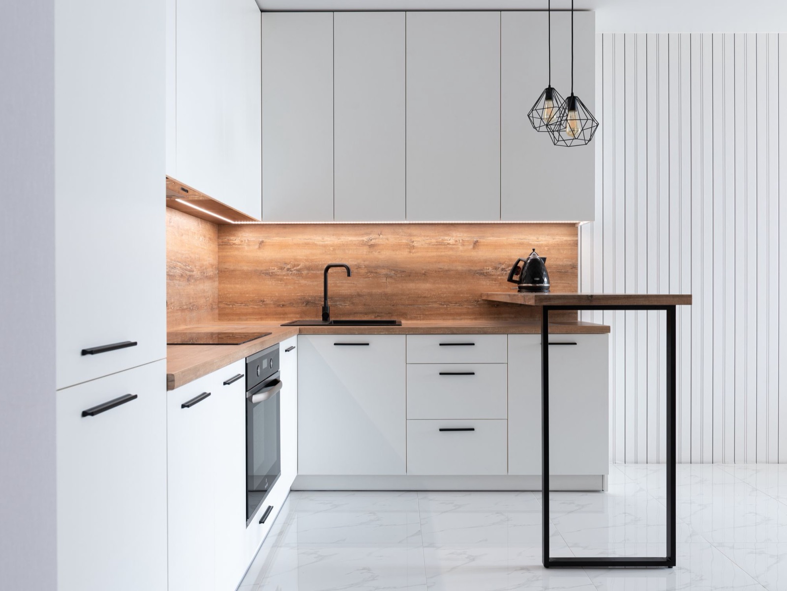 L-shaped modular kitchen with flat white matte shutters and black bar handles, a wood-look countertop and full-height backsplash lit by an LED strip under the wall units, a black sink and tap, a built-in oven, a tall unit at the left and a wood breakfast ledge on a black steel frame.
