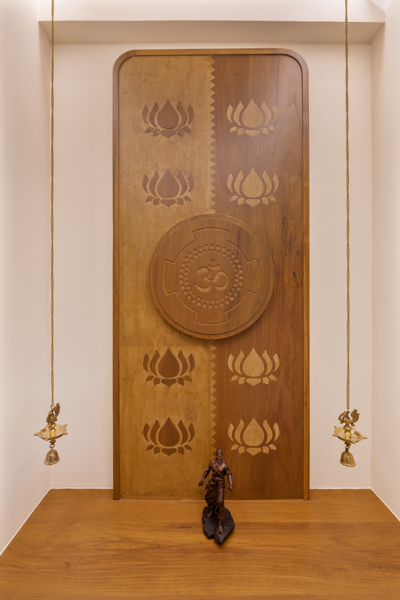 A built-in mandir in a recessed white niche: a tall wooden back panel with carved lotus motifs and a round carved Om medallion at the centre, a wooden ledge in front holding a small bronze figure, and two brass hanging lamps with bells on long chains on either side